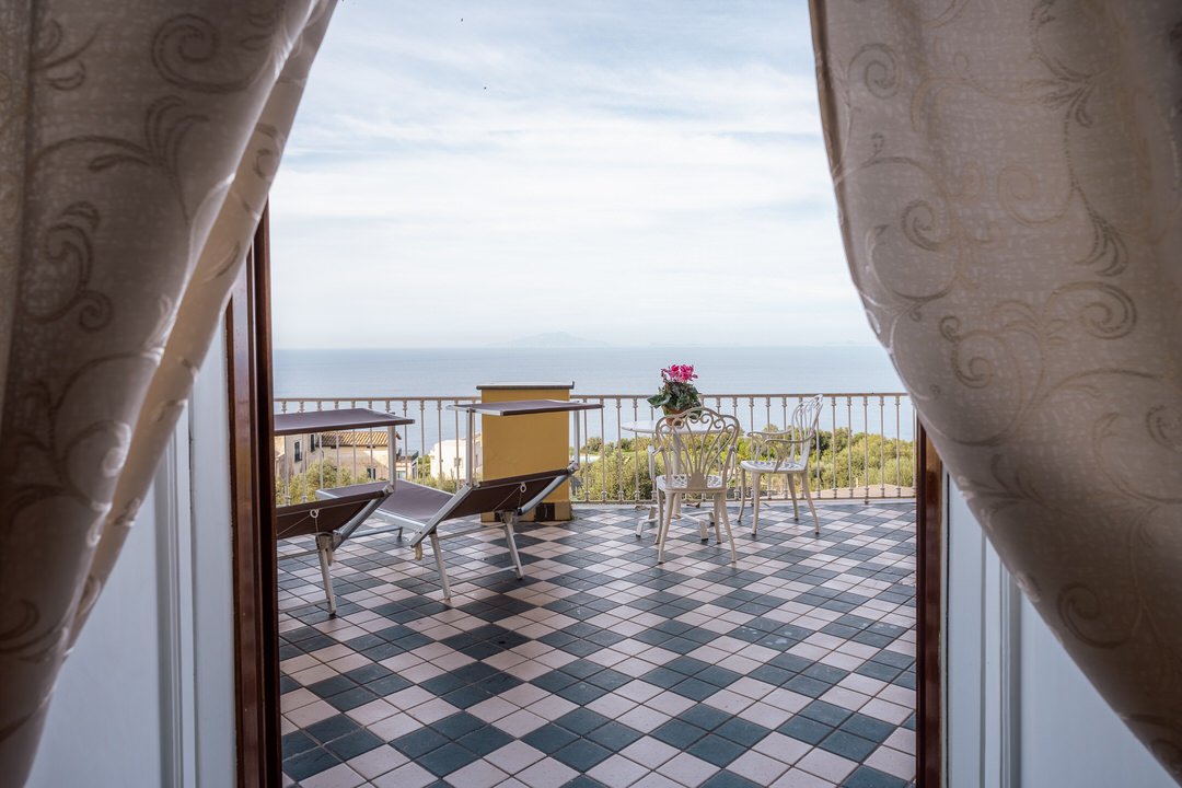 Balcony with a checkered floor, white patio furniture, potted plant with pink flowers, and a view of the sky and distant landscape is seen through open curtains.