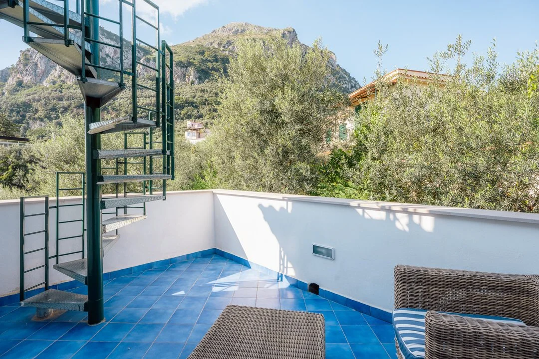 Outdoor balcony with blue tiled flooring, wicker furniture, a white wall, and green trees with mountain in the background.
