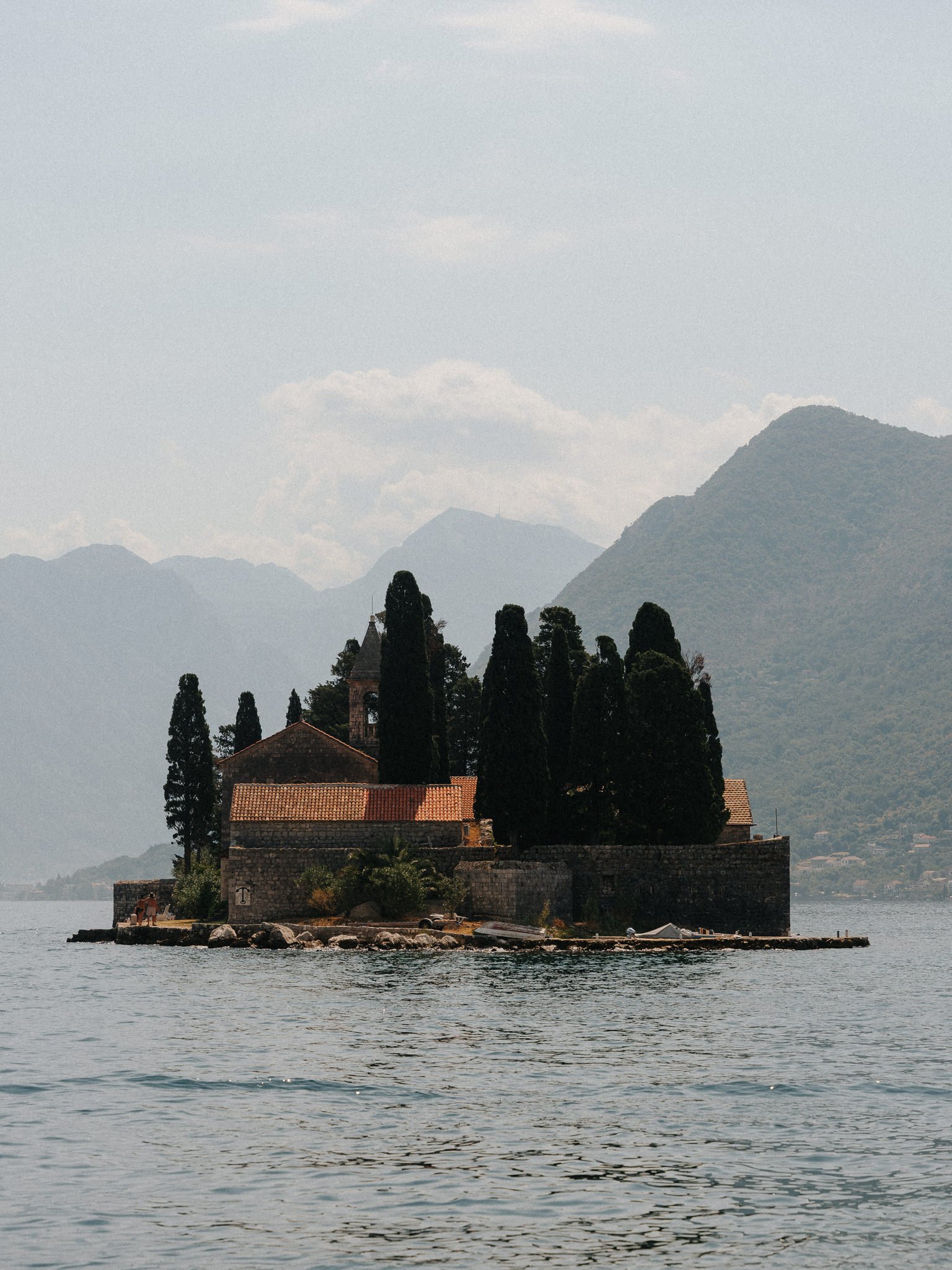 A small island with a church and tall trees is in a body of water, with mountains and cloudy sky in the background.