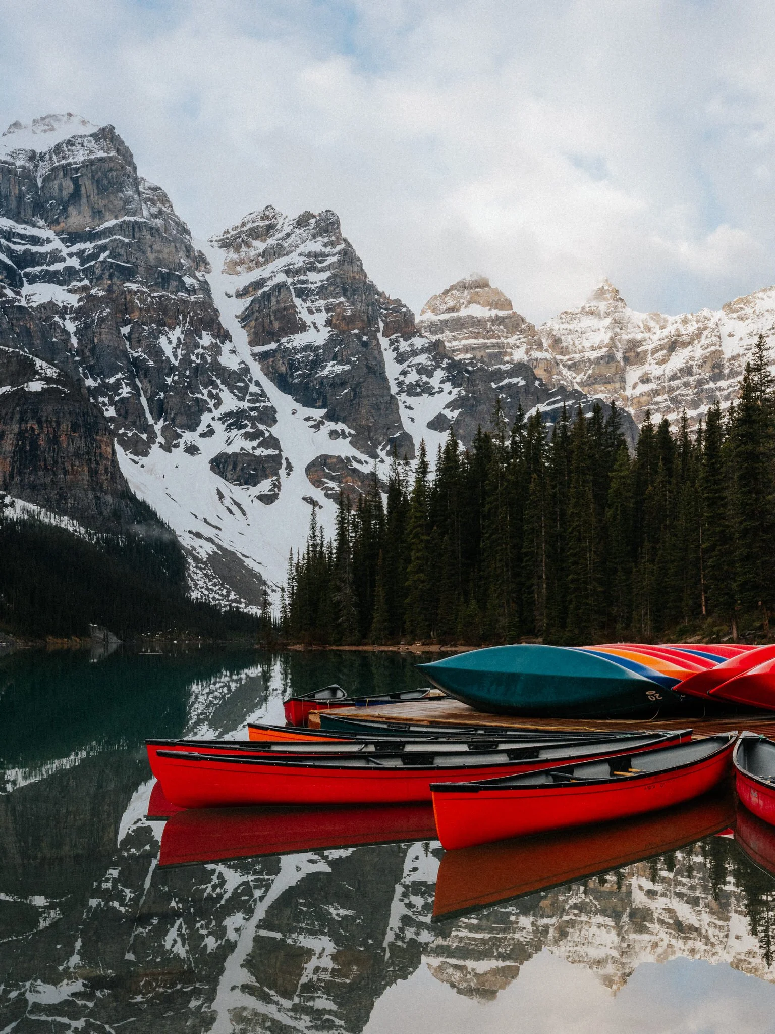 Colorful kayaks on calm mountain lake with snow-covered peaks and dense forest in the background.