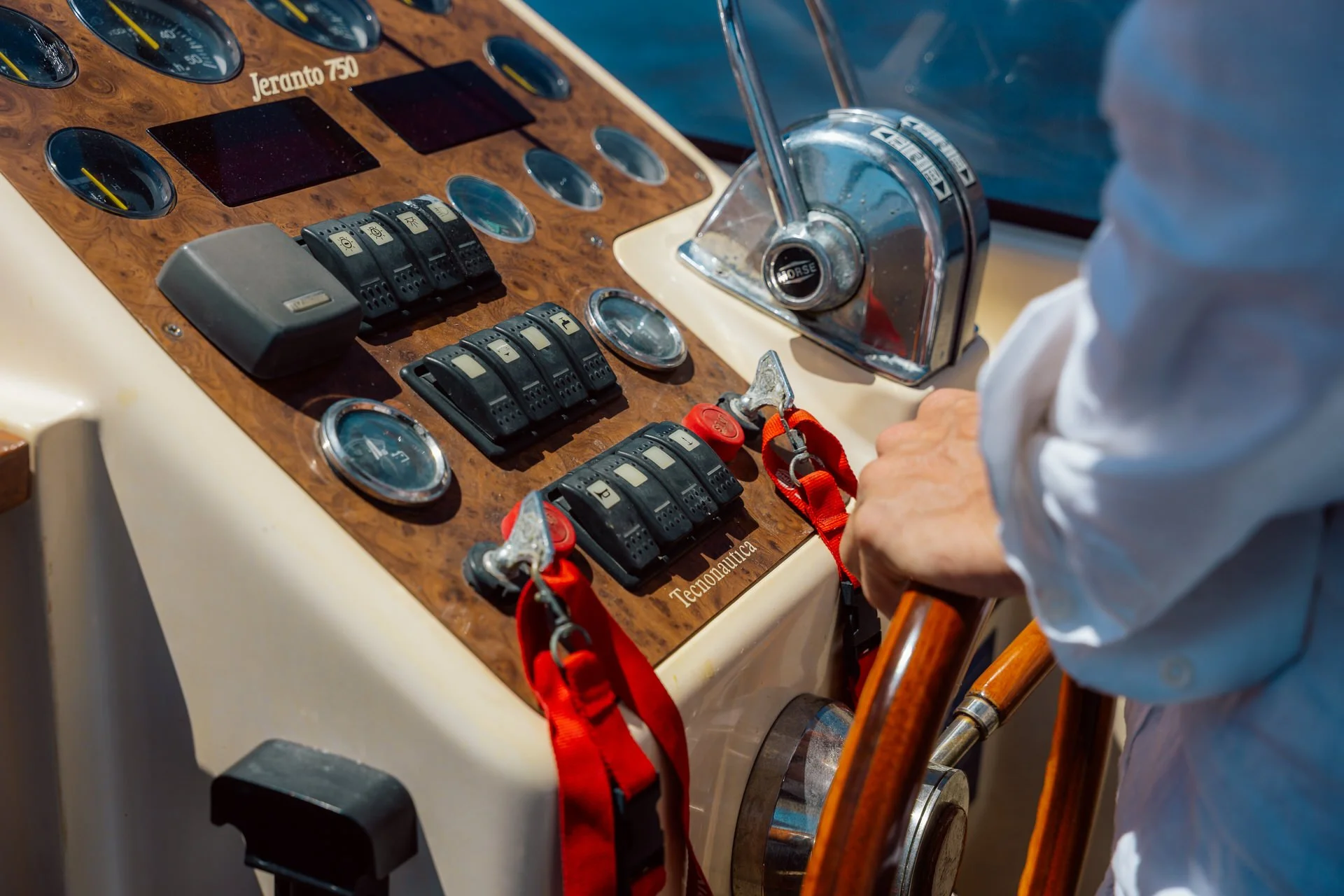Close-up of a yacht's wooden control panel with various gauges, switches, and a throttle lever, with a person's hand on the steering wheel.