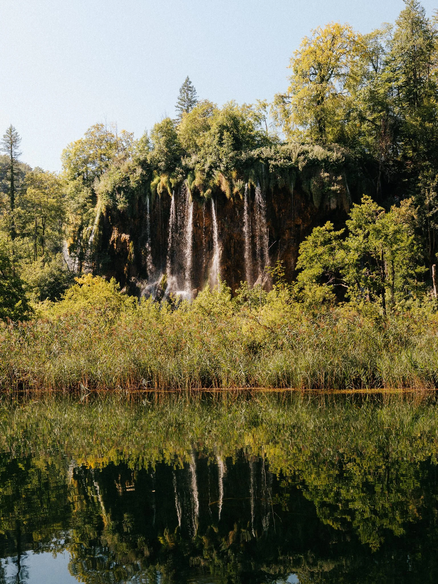 A serene landscape of a multi-tiered waterfall cascading down a rocky cliff surrounded by green trees, reflected in a calm body of water below.