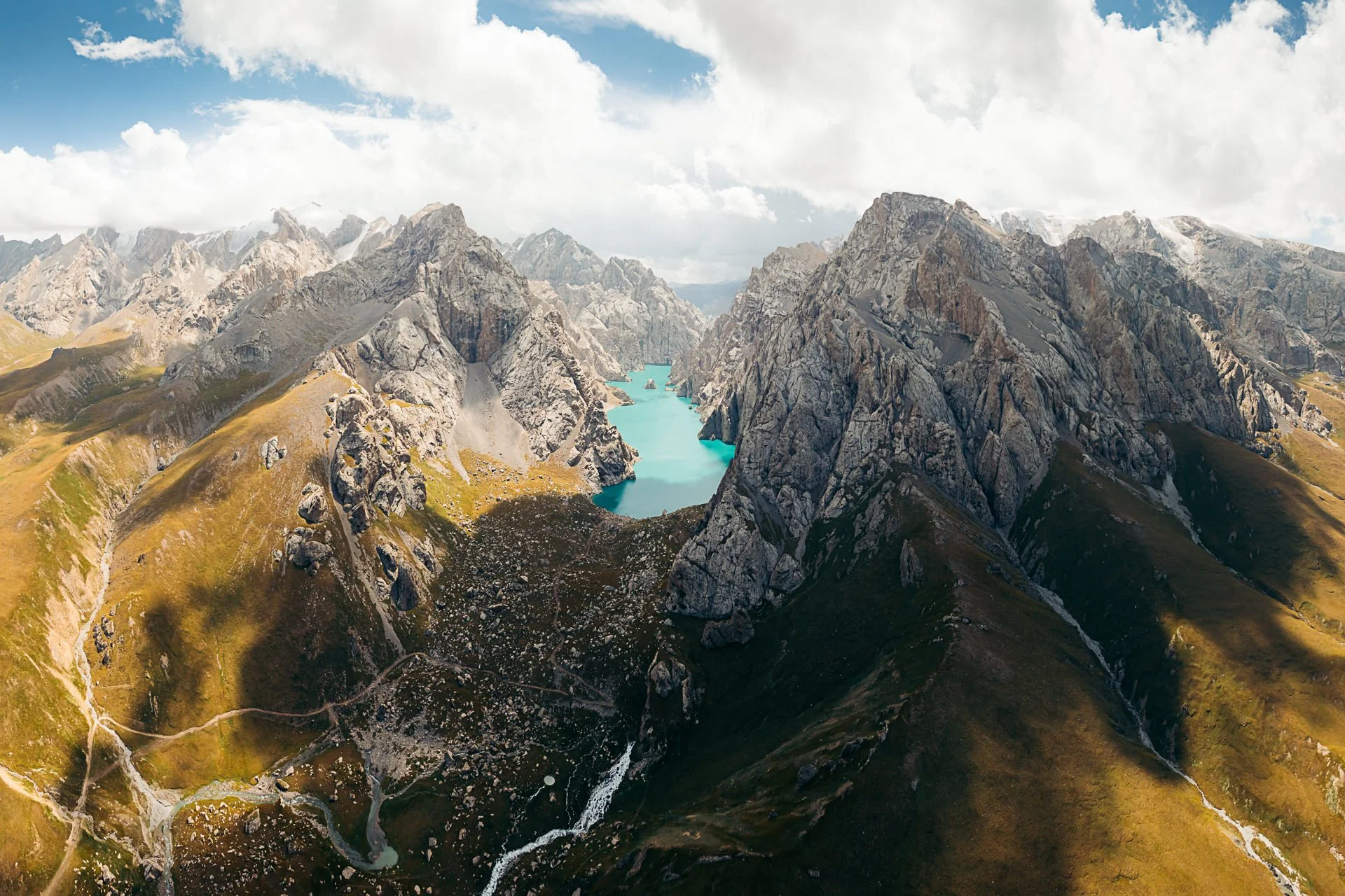 Aerial view of a mountain range with a turquoise lake in a valley surrounded by rugged peaks and green slopes.