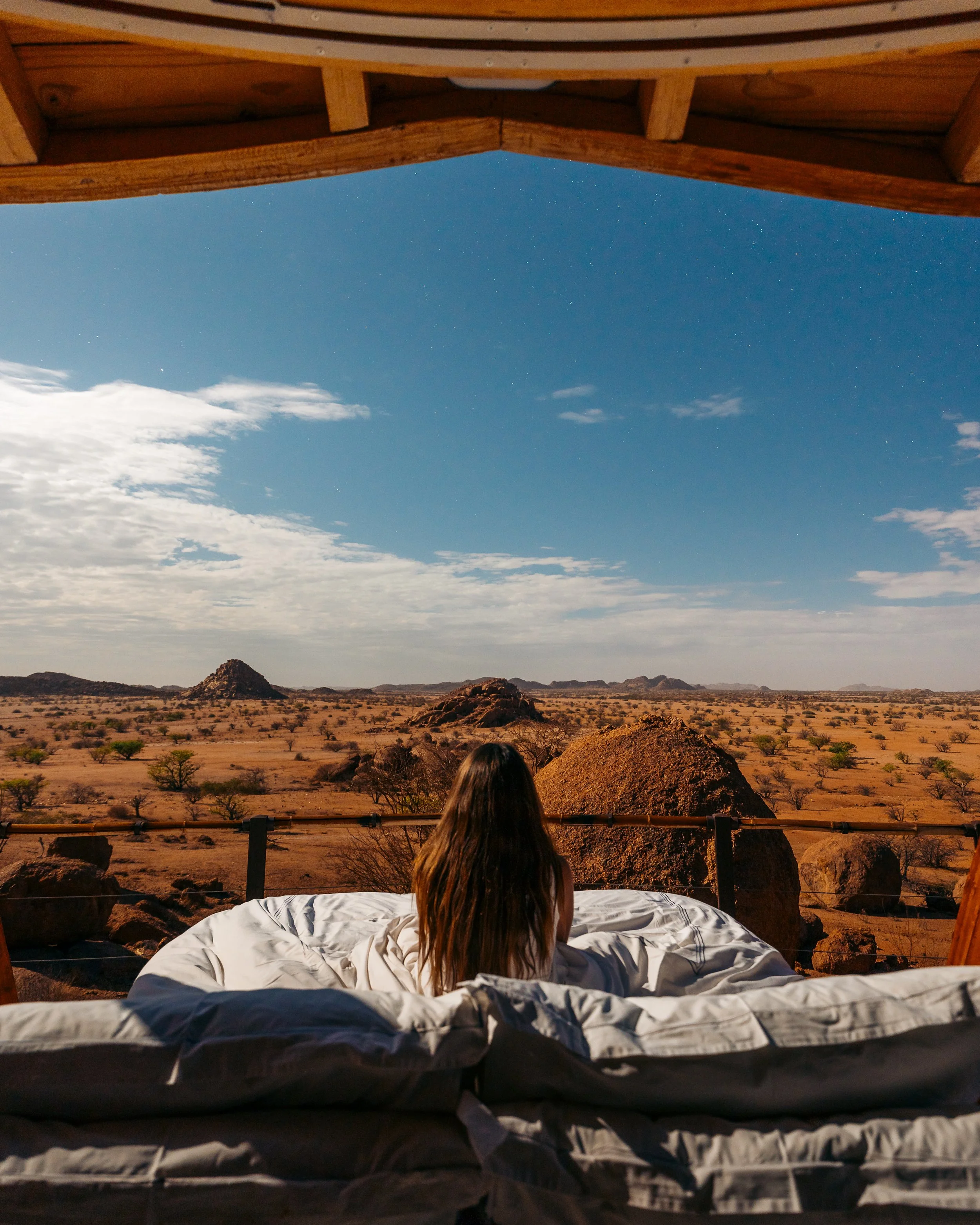 A woman with long brown hair sitting on a bed inside a wooden structure, looking out at a desert landscape with scattered bushes, large rocks, and distant mountains under a partly cloudy sky.