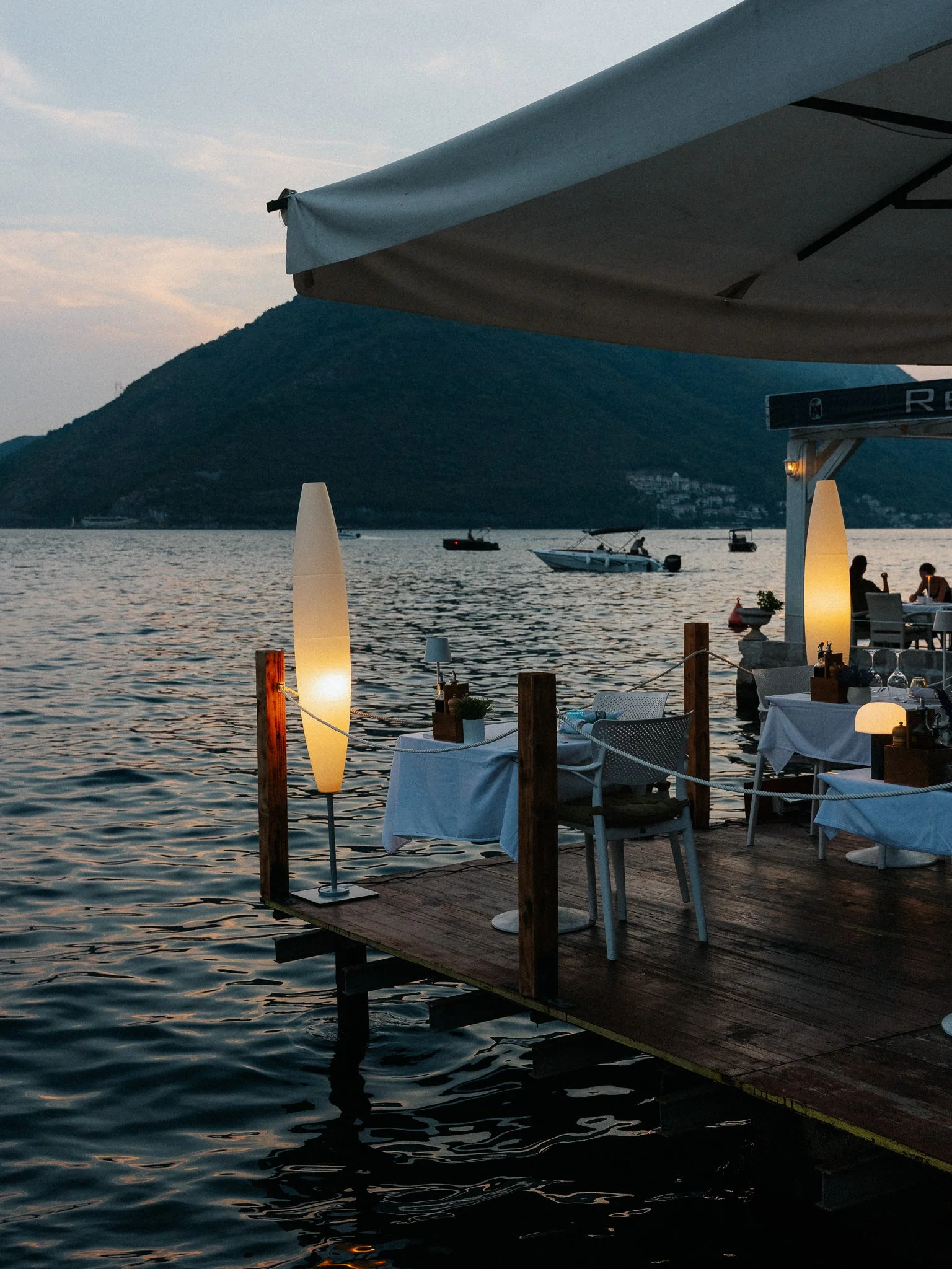 A waterfront restaurant or cafe at sunset with tables set with white tablecloths, chairs, lamps, and small potted plants. Boats are visible on the water, and a mountain in the background.