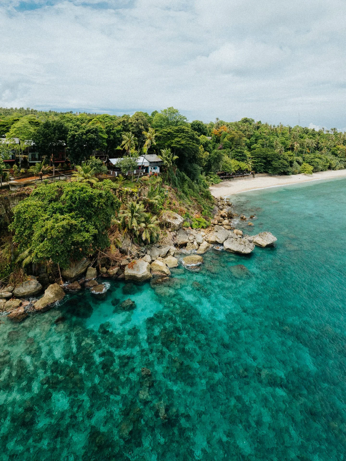 A coastal scene with turquoise water, rocky shoreline, and lush green trees with some houses on the hill.