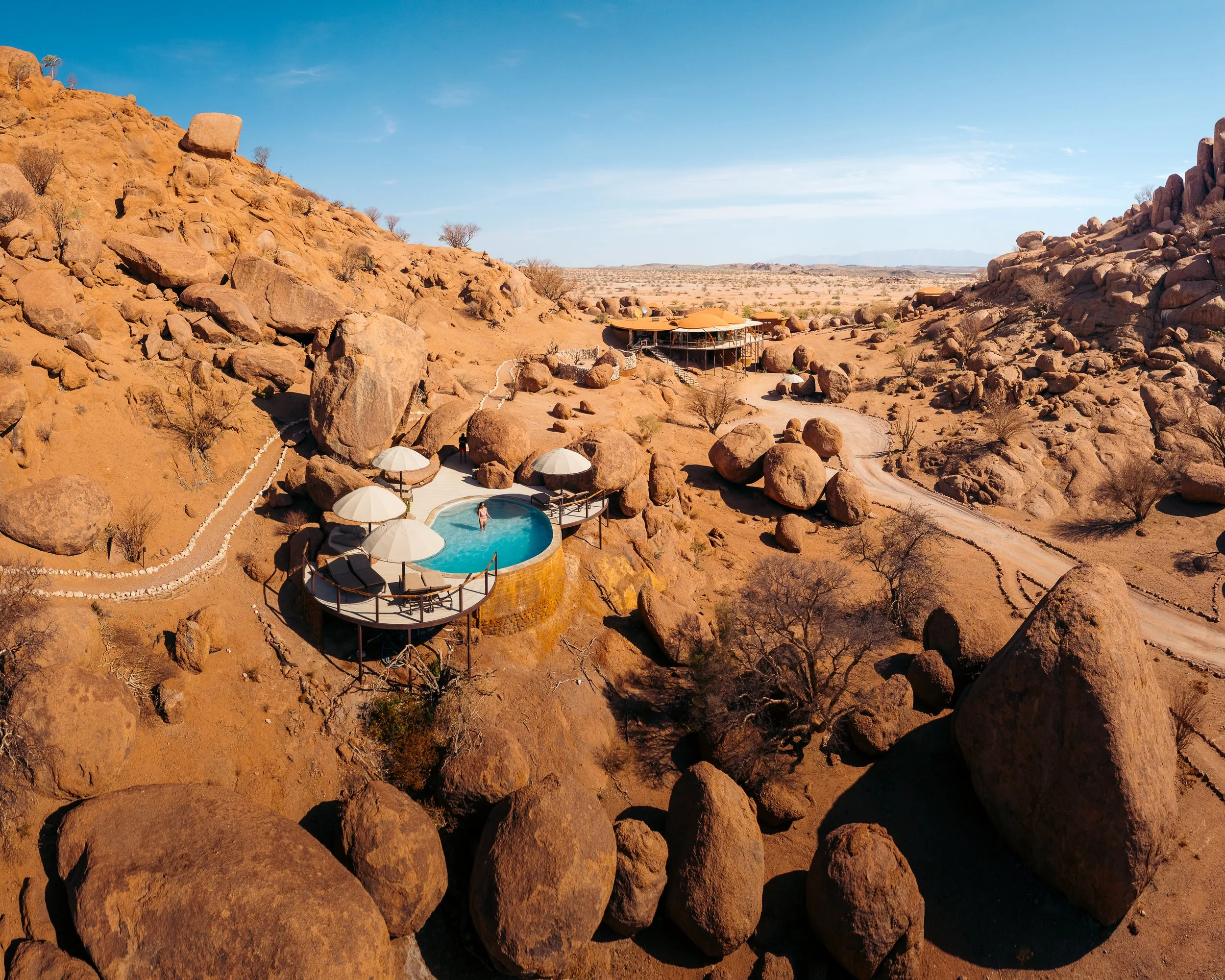 A desert landscape with large rocks and a modern building and pool on a hill. The sky is clear and blue.