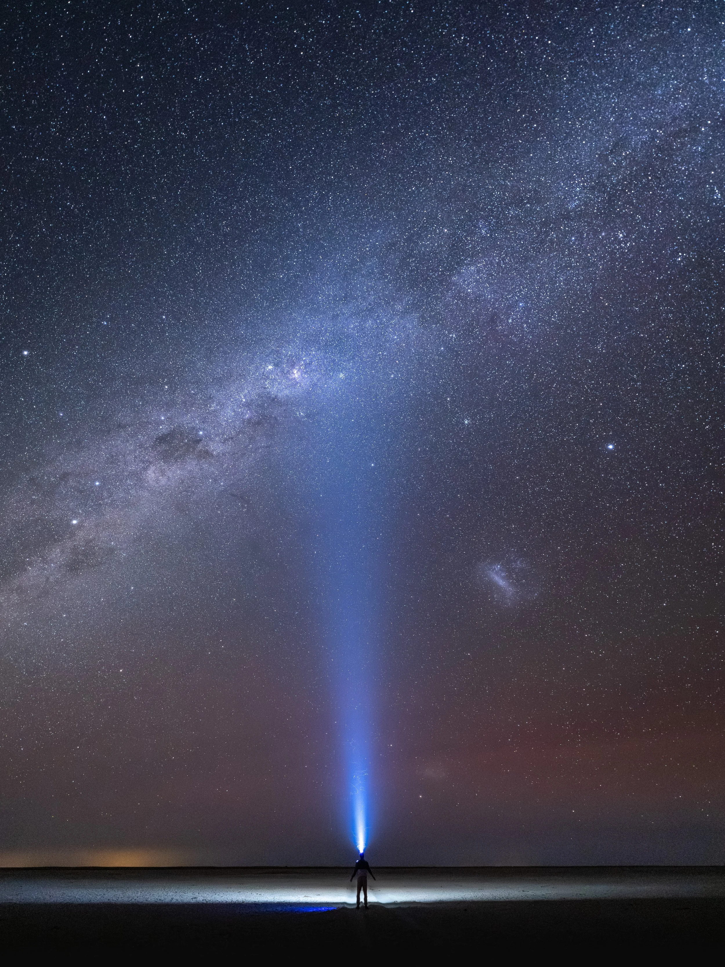 A person on a beach pointing a blue laser beam toward the sky, which displays a starry night with the Milky Way galaxy visible across the sky.