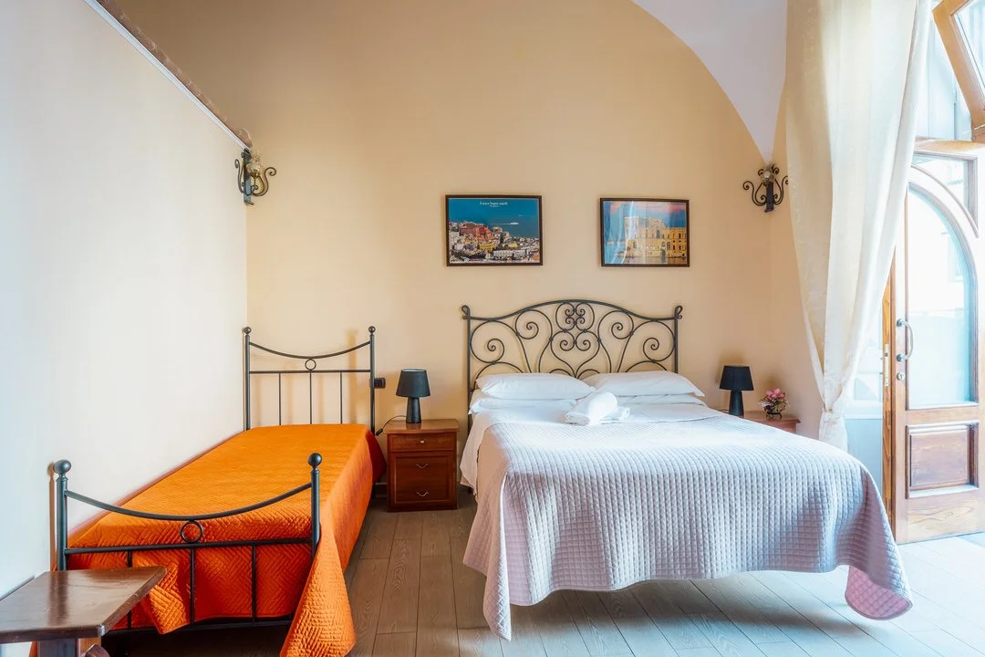 A bedroom with a metal bed frame, white bedding, and a wooden nightstand with a black lamp. There is a single bed with an orange cover and a small side table. The room has beige walls, two framed pictures above the bed, a wooden door with a window, a