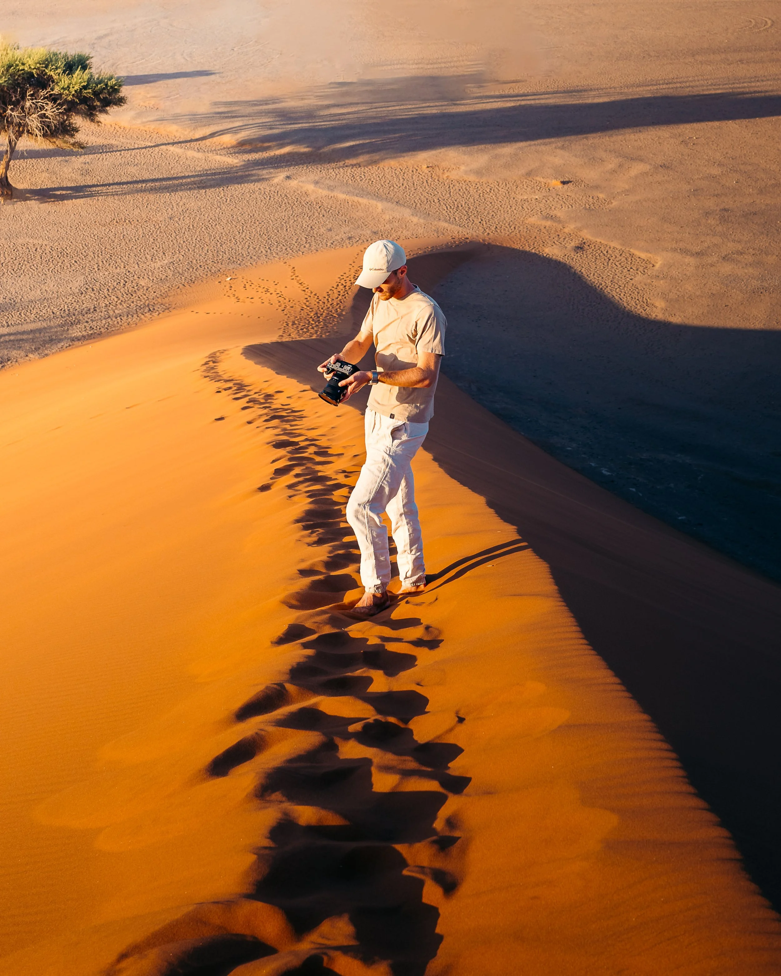 A man standing on a sand dune in a desert during sunset, holding a camera and looking down at it.