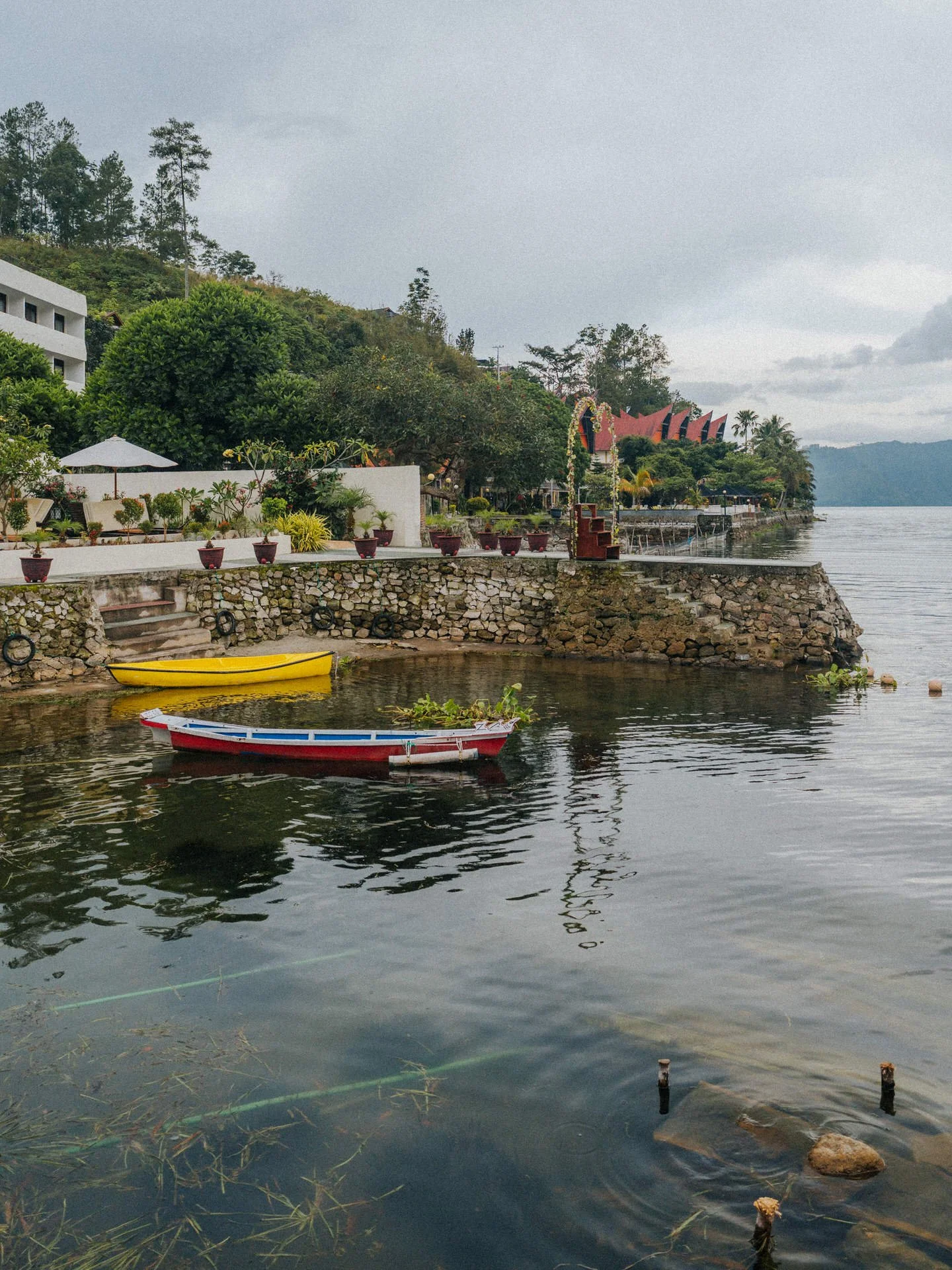 A scenic lakeside with boats, a stone wall, lush greenery, outdoor seating, and buildings on a cloudy day.