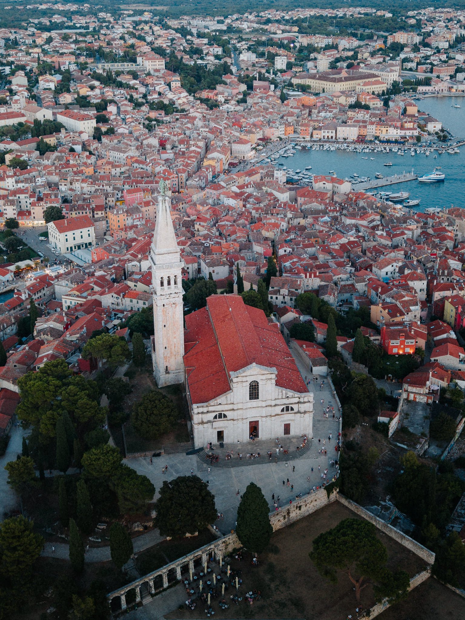 An aerial view of a coastal town with a large church in the foreground and a marina with boats in the background. The town has a dense cluster of buildings with red-tiled roofs, trees, and streets.