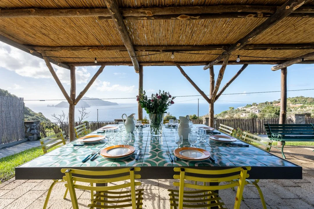 Outdoor dining table set with plates, glasses, and cutlery, under a thatched roof with a scenic ocean view in the background.