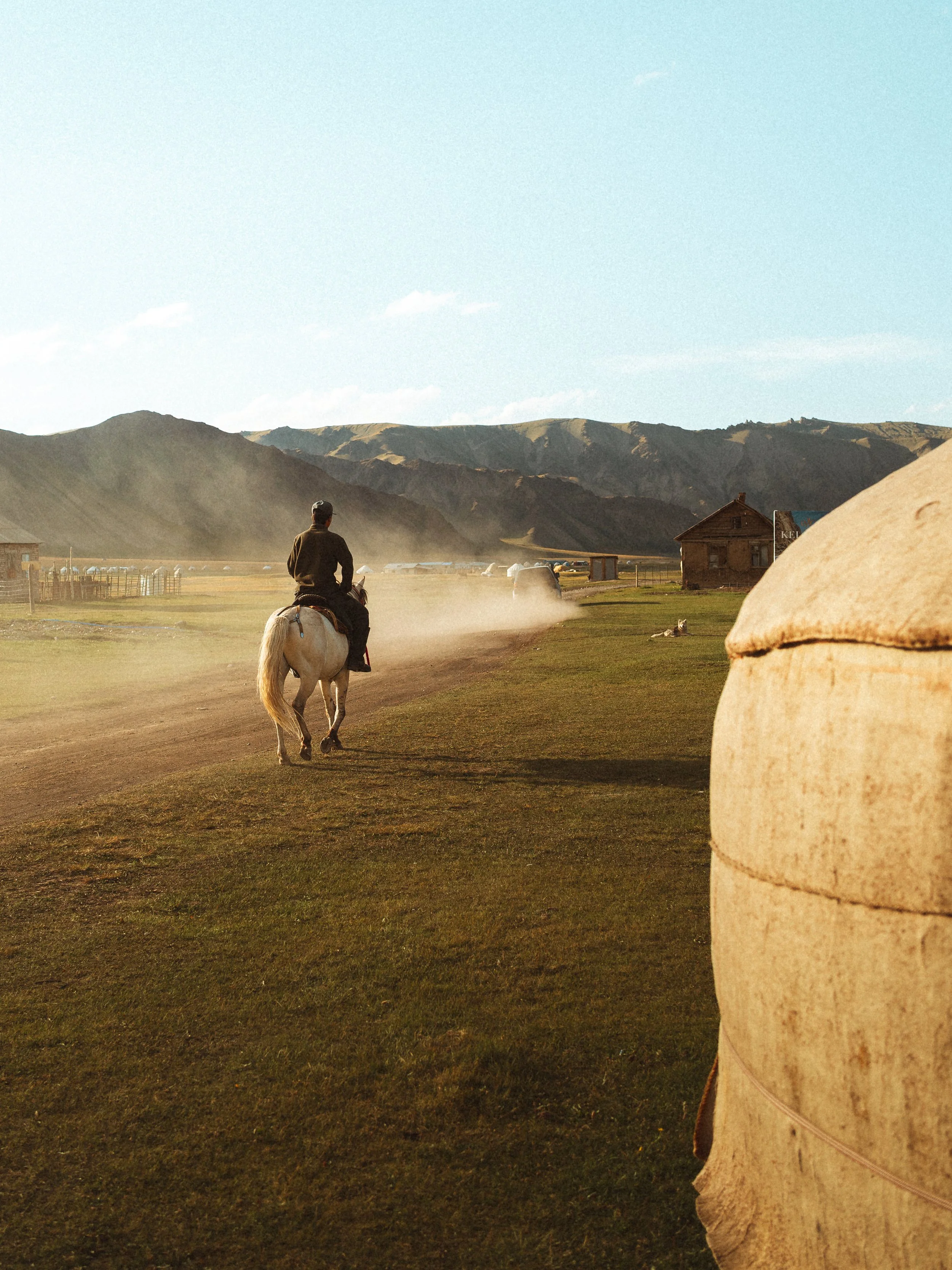 A person riding a white horse on a dirt path in a rural area with mountains in the background