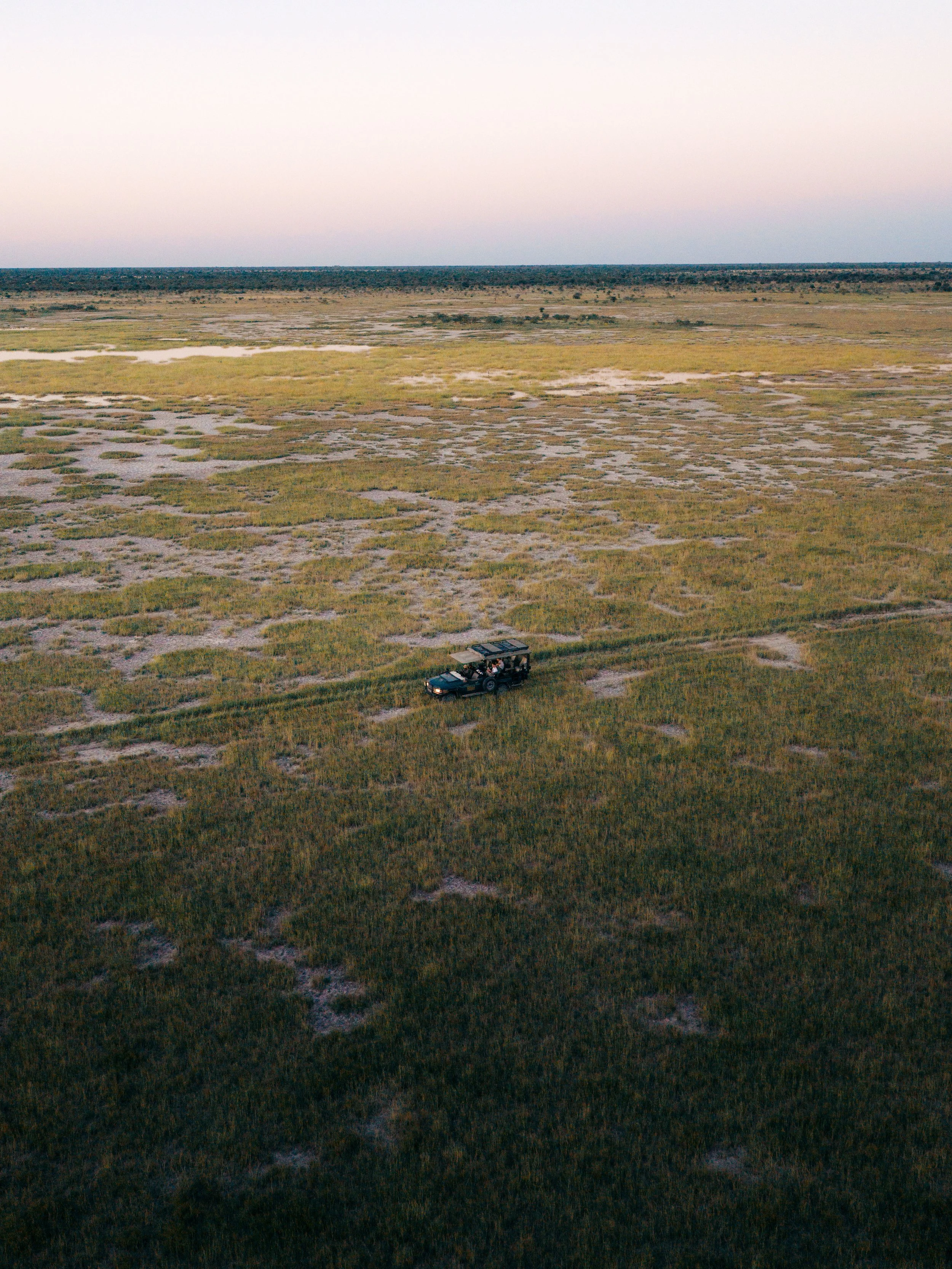 A vast, flat landscape with patches of grass and small water pools, with a single safari vehicle carrying passengers in the foreground, and a distant horizon with a sky that is transitioning from sunset to dusk.