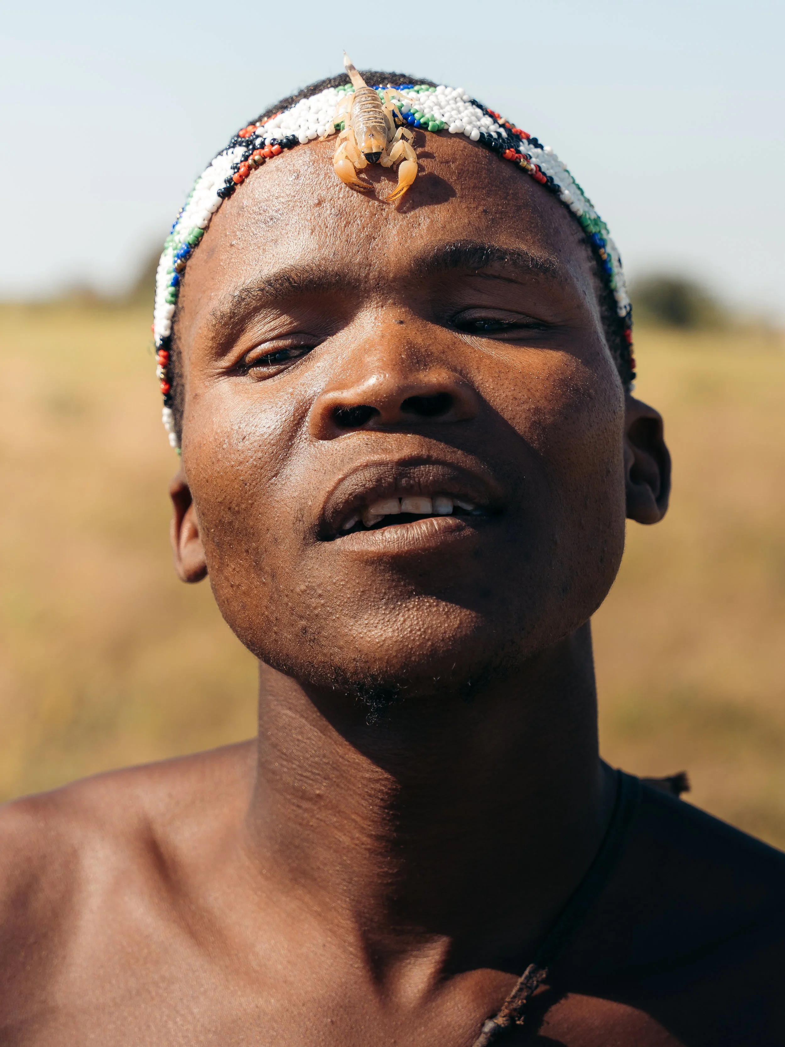 A person with dark skin and closed eyes wearing a beaded headband decorated with colorful beads and a small animal figurine, outdoors with a blurred landscape background.