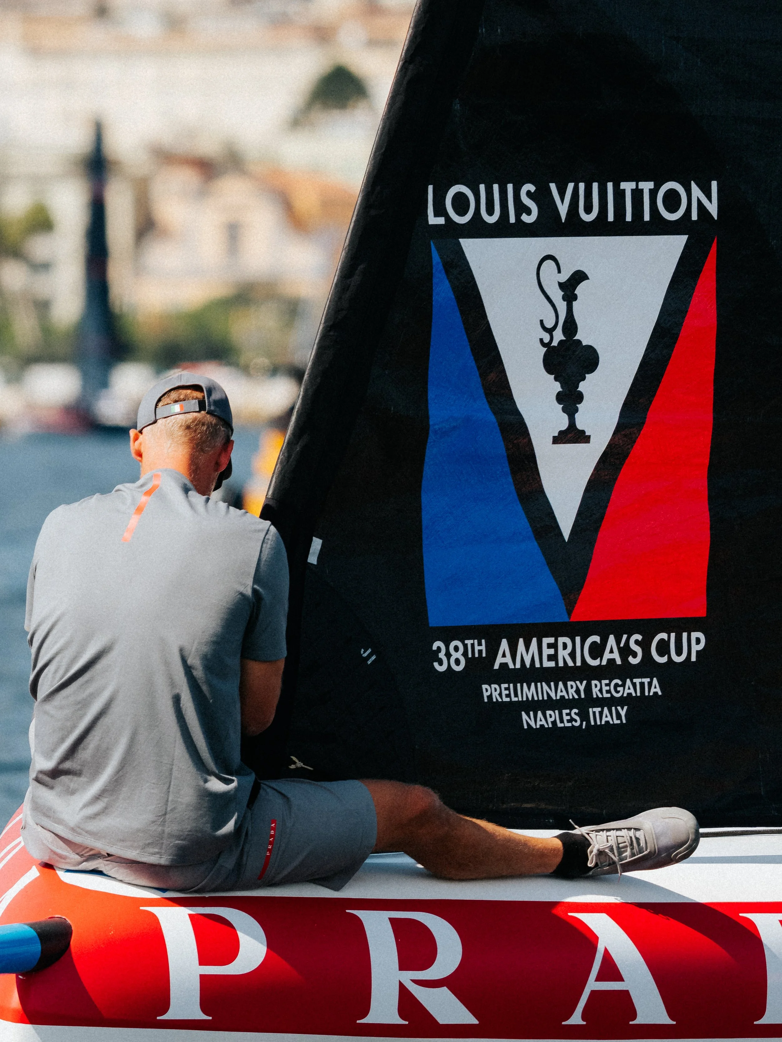 A person wearing a gray cap, gray shirt, blue shorts, and sneakers seated on a boat, with a large black sail displaying the logo for the 38th America's Cup in Naples, Italy.