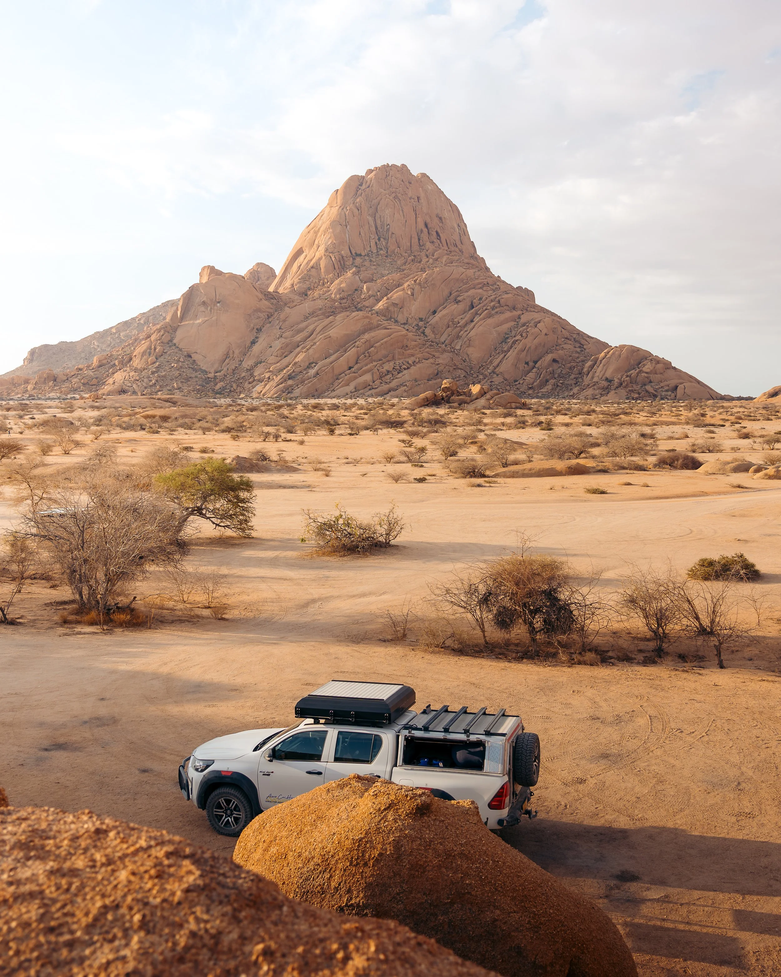 A white off-road vehicle parked in a desert with sparse vegetation and a large rocky hill or mountain in the background.