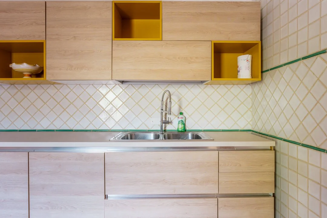 Empty kitchen sink with a green soap bottle, wooden cabinets above and below, beige tiled wall with green grout lines.