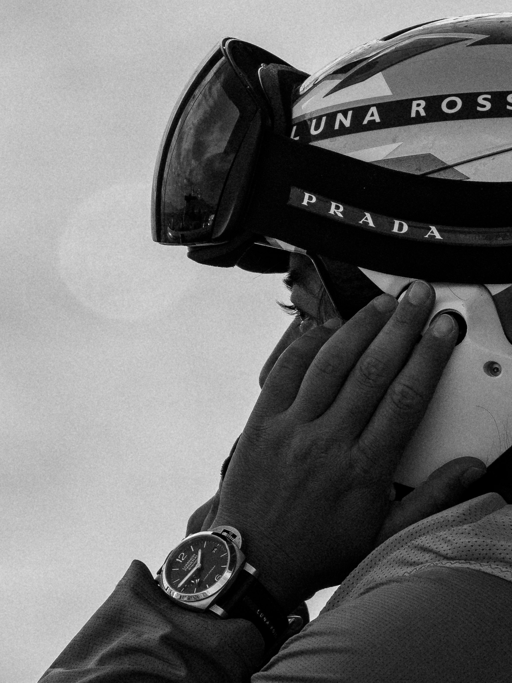 A person wearing dark athletic clothing and a watch, holding a helmet with their hand, with a background of clear sky.