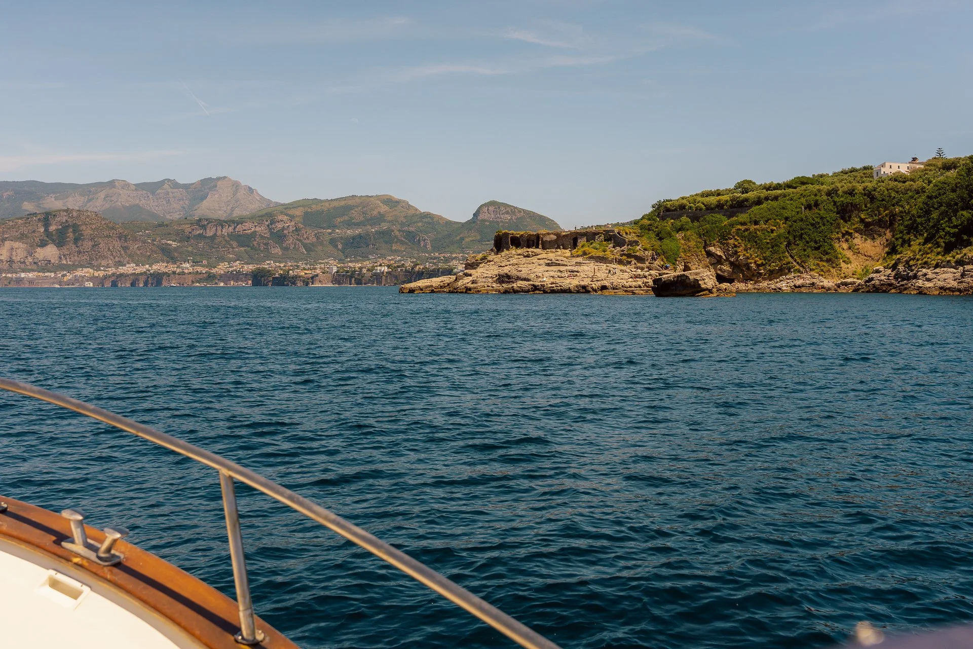 View of the coastline with rocky cliffs, green vegetation, and mountains in the distance, seen from a boat on calm blue water.