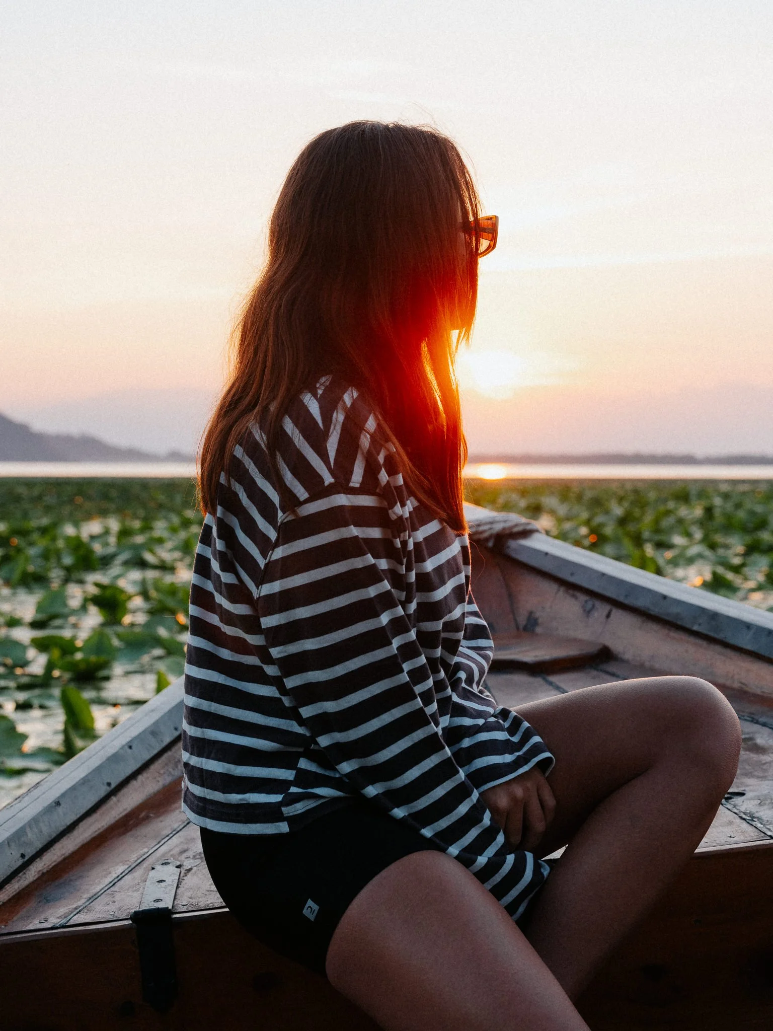 A woman with long hair and glasses sitting in a boat during sunset, with water and distant hills in the background.