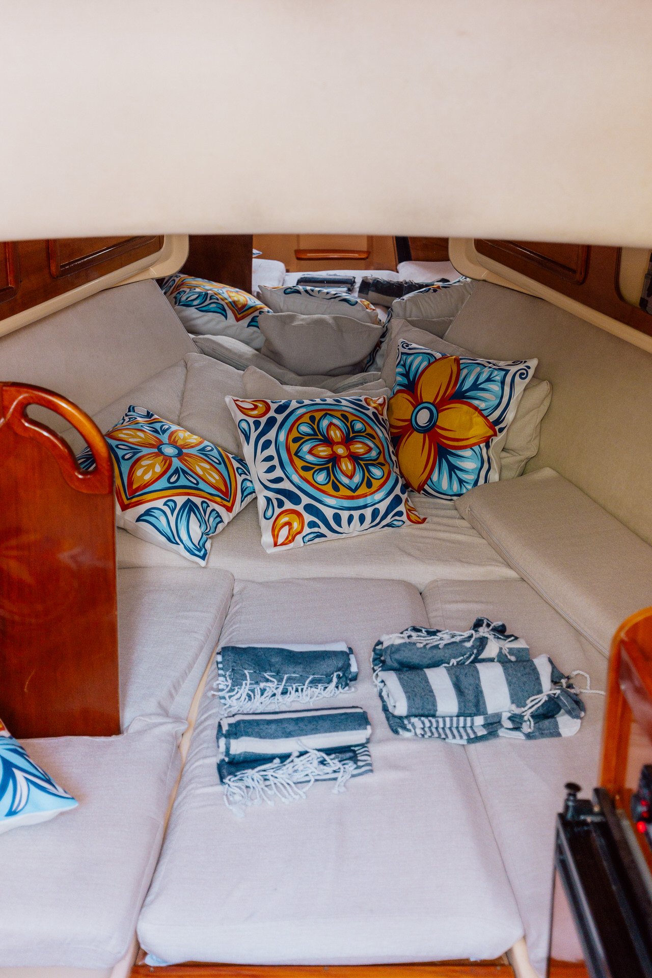 Inside a boat's cabin with a beige sectional sofa covered with colorful floral and blue patterned pillows, and stacked striped and patterned towels, some visible in the foreground.