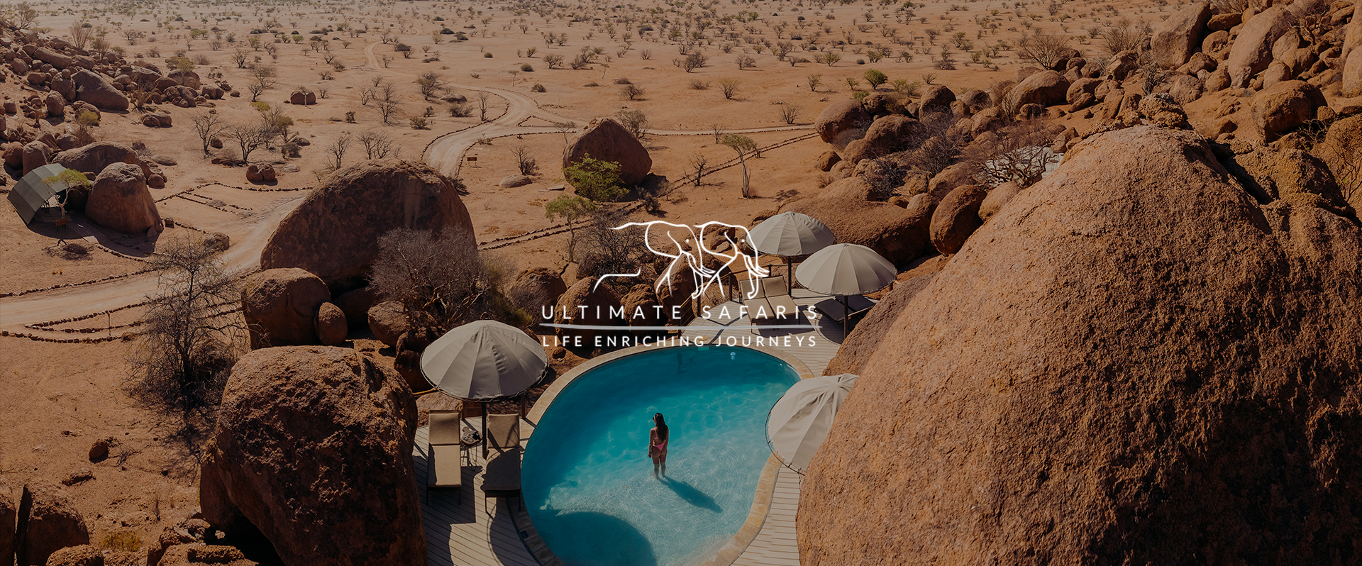 A woman standing in a swimming pool in a desert landscape with large rocks and sparse trees, with umbrellas and lounge chairs nearby, and the logo and slogan of Ultimate Safaris. 