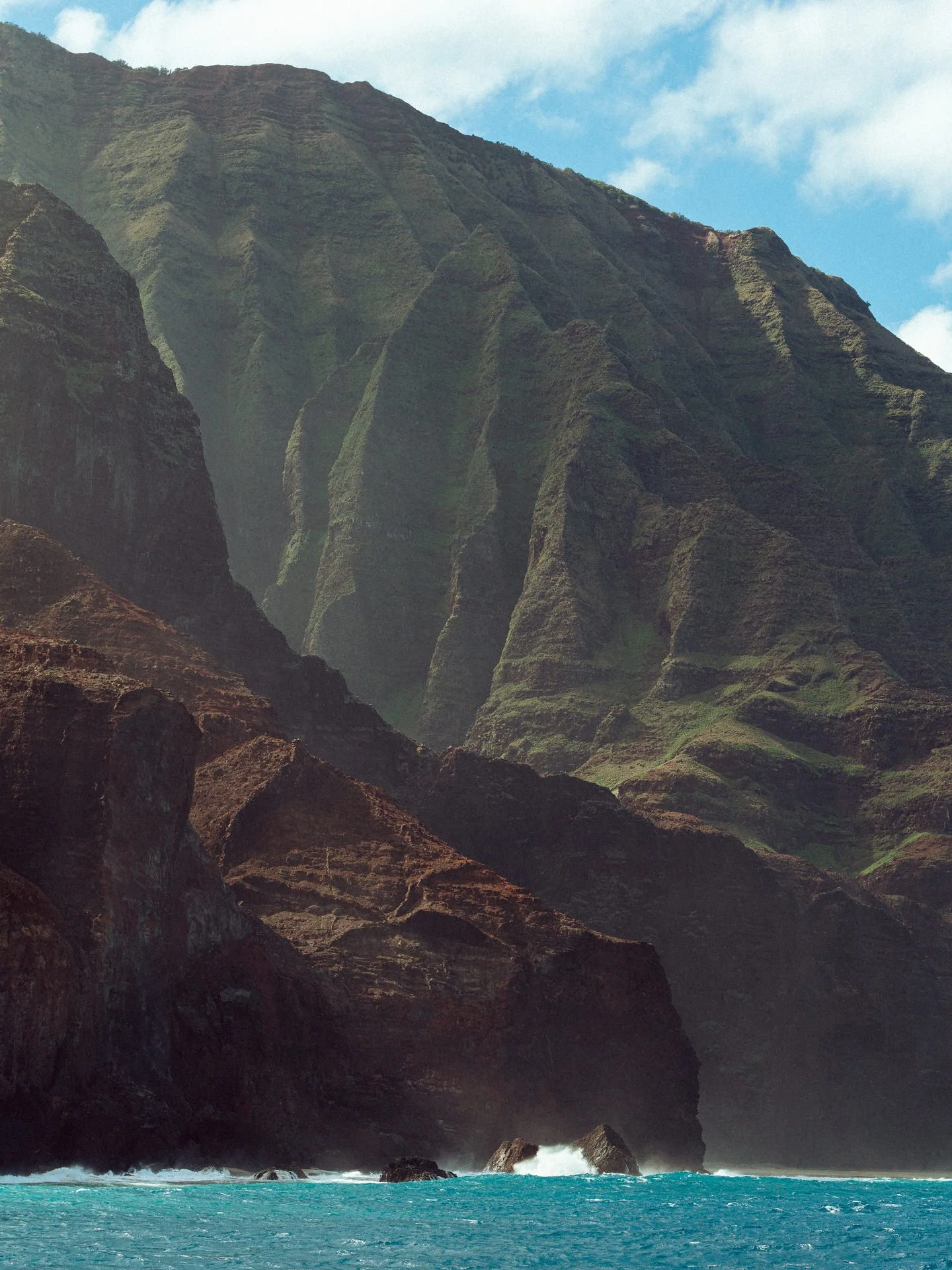 Cliffside ocean view with steep, lush green mountains and a blue sky with clouds.