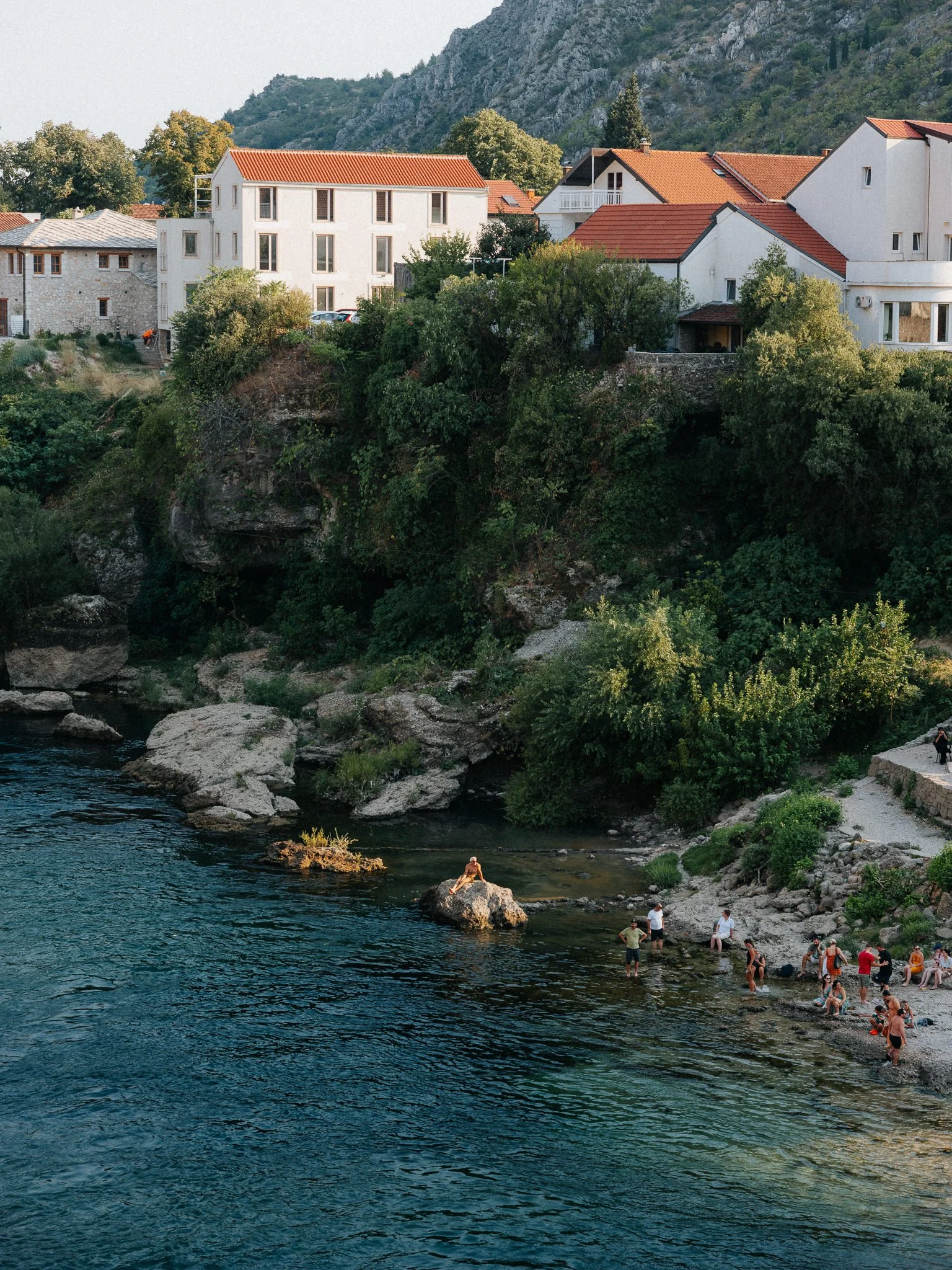 People swimming and relaxing at a rocky riverside with green trees and white houses with orange roofs in the background.