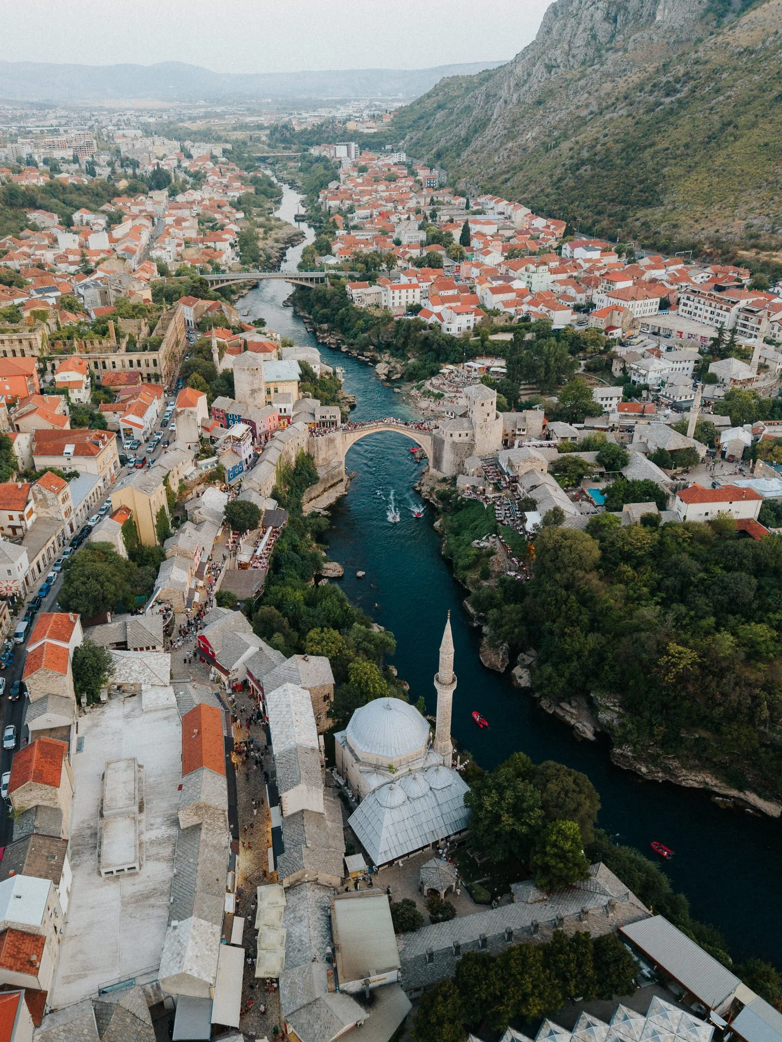 Aerial view of a historic city with a river running through it, featuring bridges, white buildings with red-tiled roofs, and a minaret surrounded by green trees, with mountains in the background.