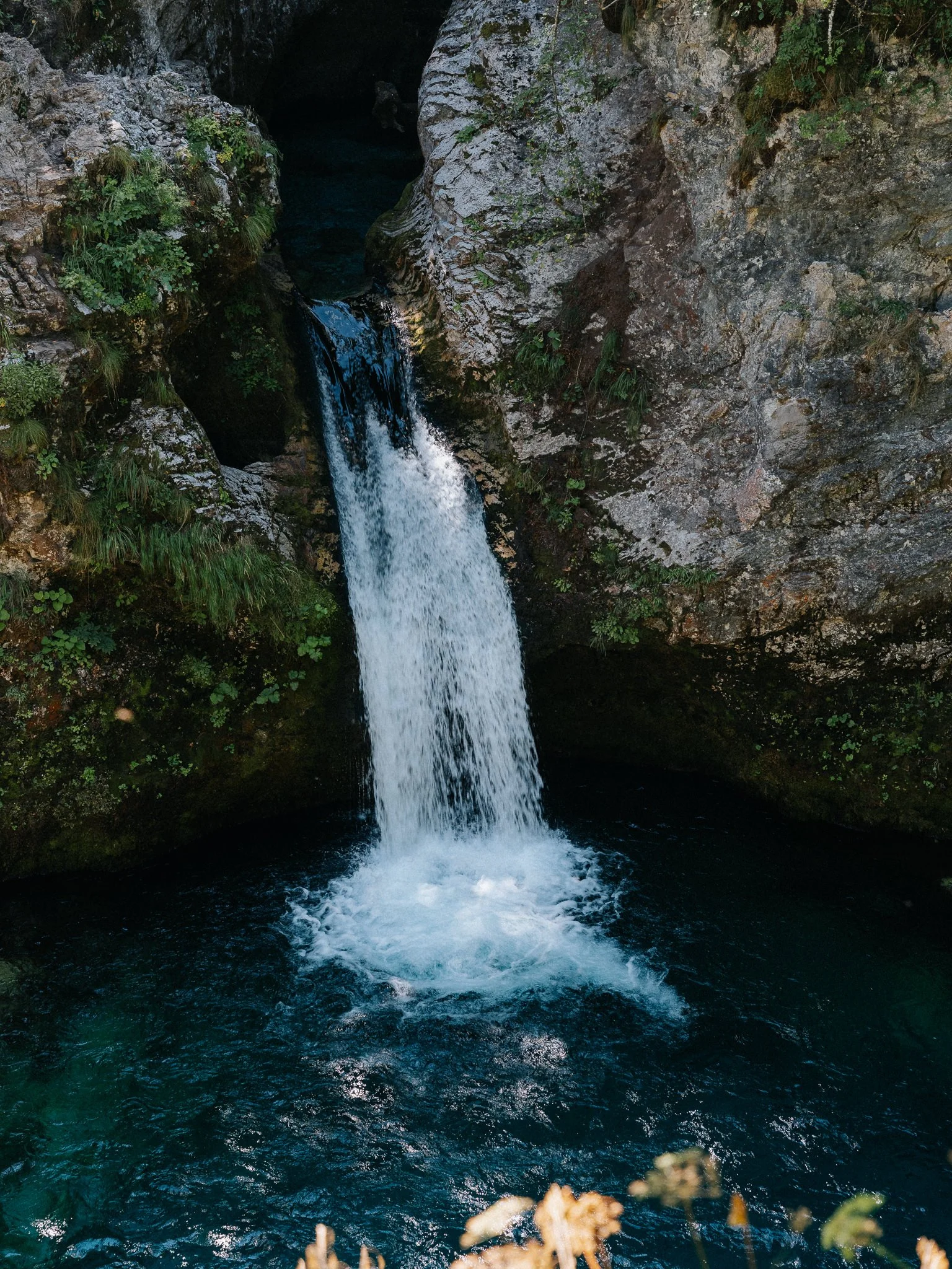 A waterfall flowing into a dark pool of water surrounded by rocky cliffs and green vegetation.