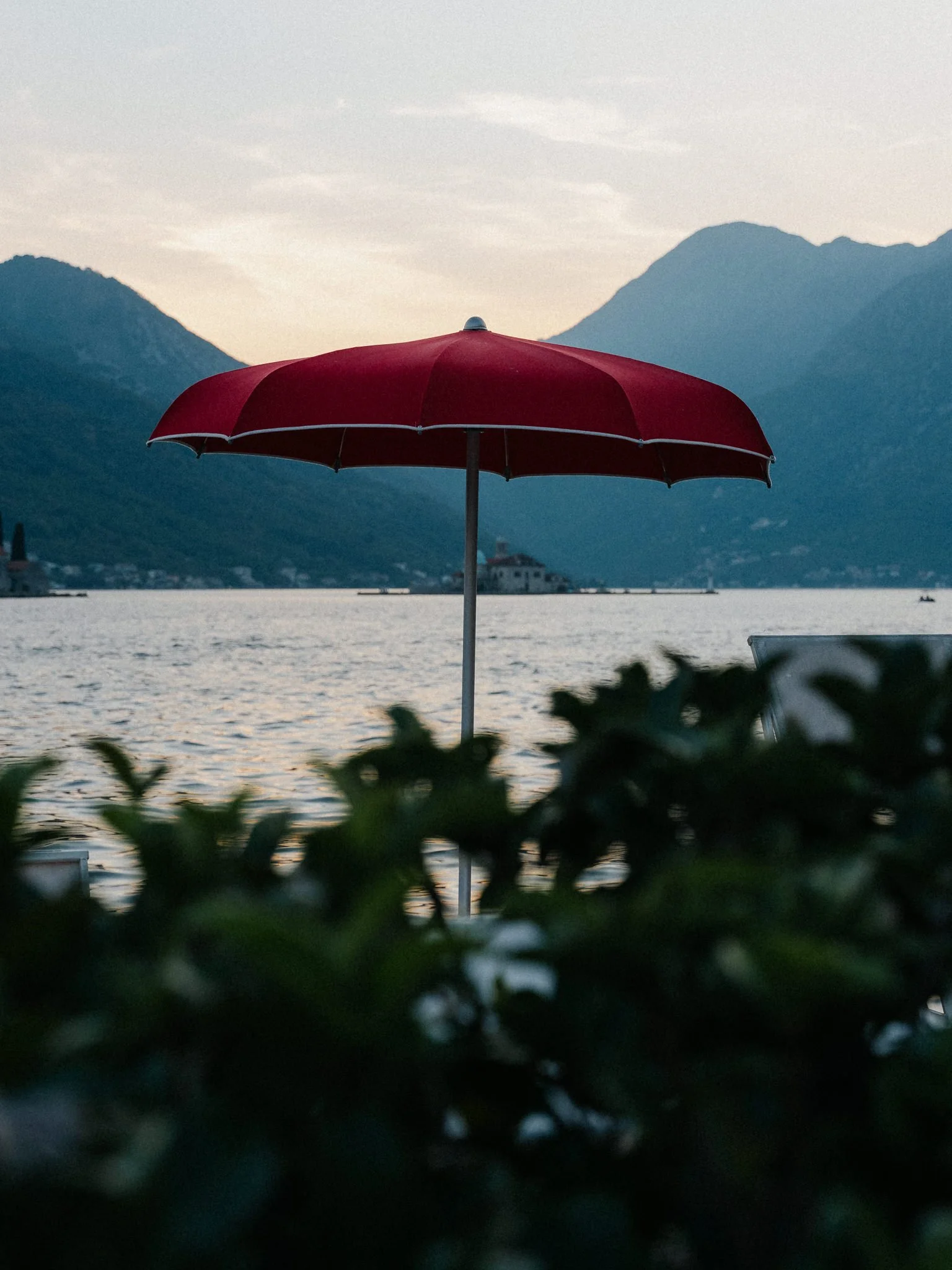 A red patio umbrella stands open by a lakeside with mountains in the background, near a house and a boat on the water during dusk.