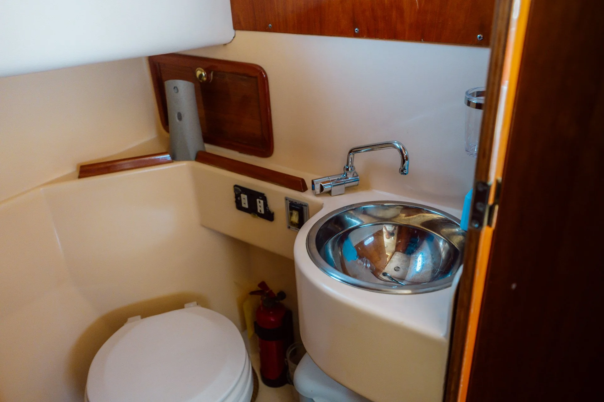 Small boat bathroom with a toilet, a stainless steel sink, a wall-mounted faucet, wooden cabinet, and electrical outlets.