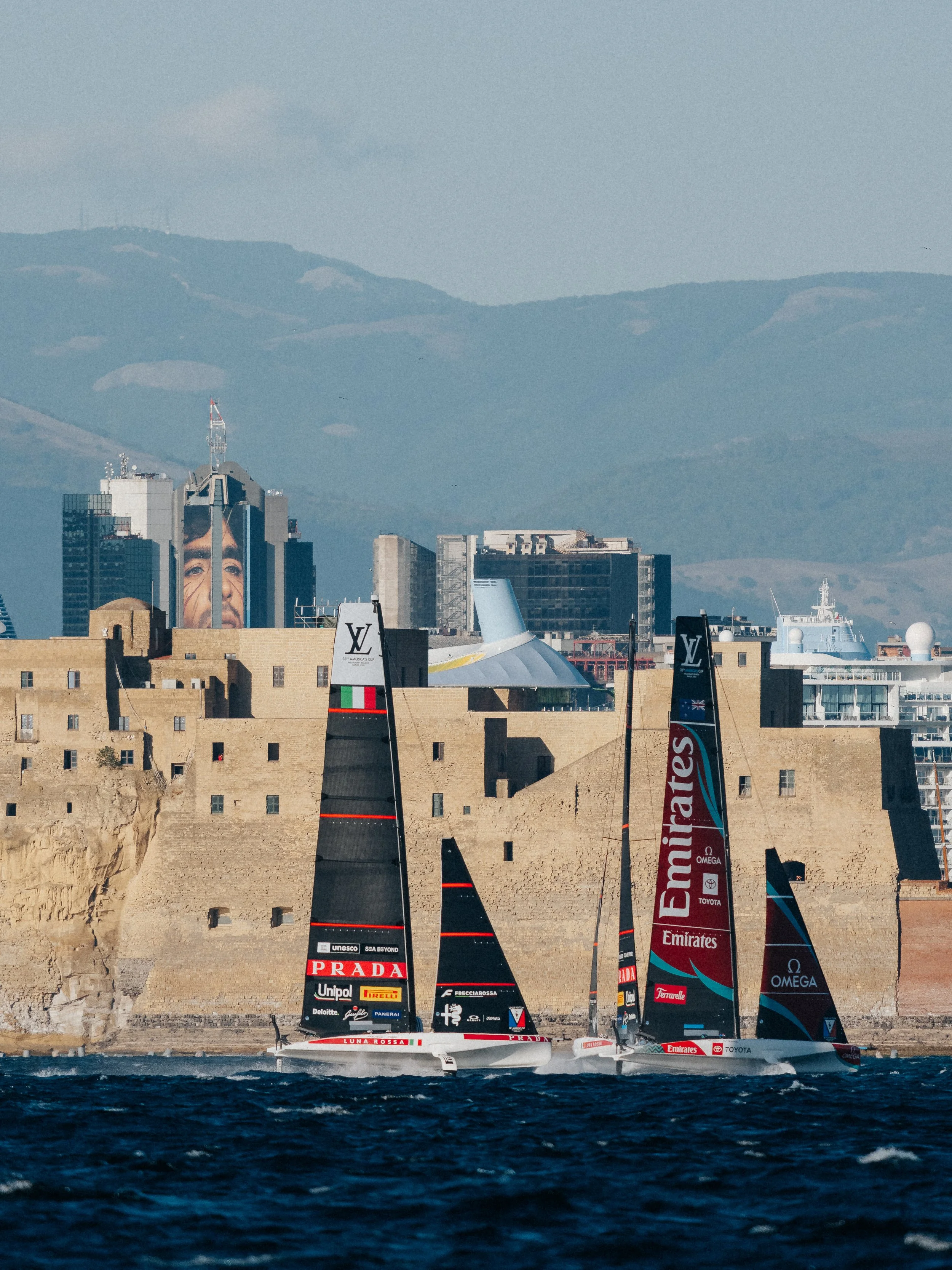 Two high-tech racing sailboats with large black and red sails racing on the water, with a cityscape and distant mountain range in the background.