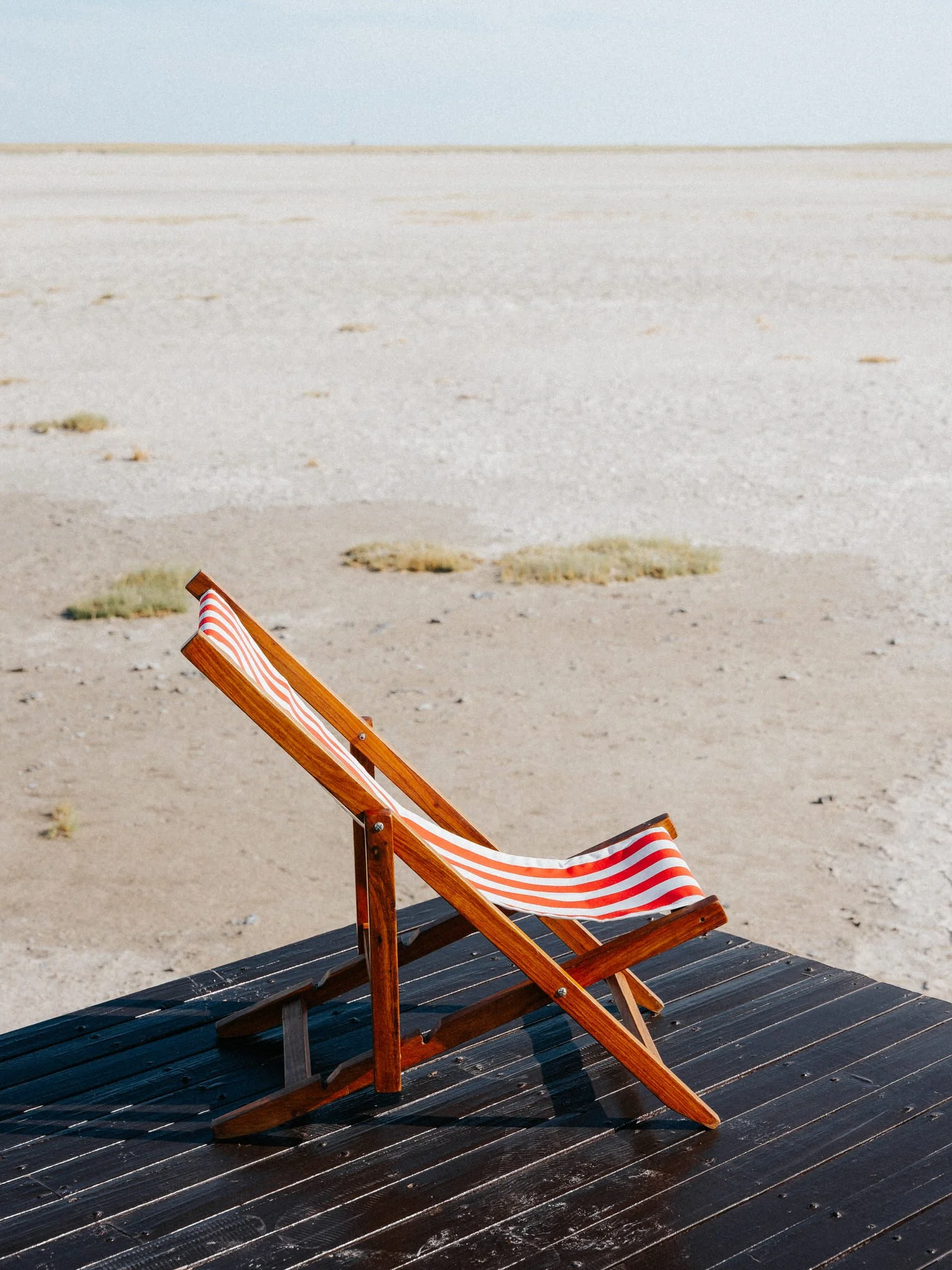 A striped orange and white beach chair on a black wooden deck, with a sandy beach and a cloudy sky in the background.