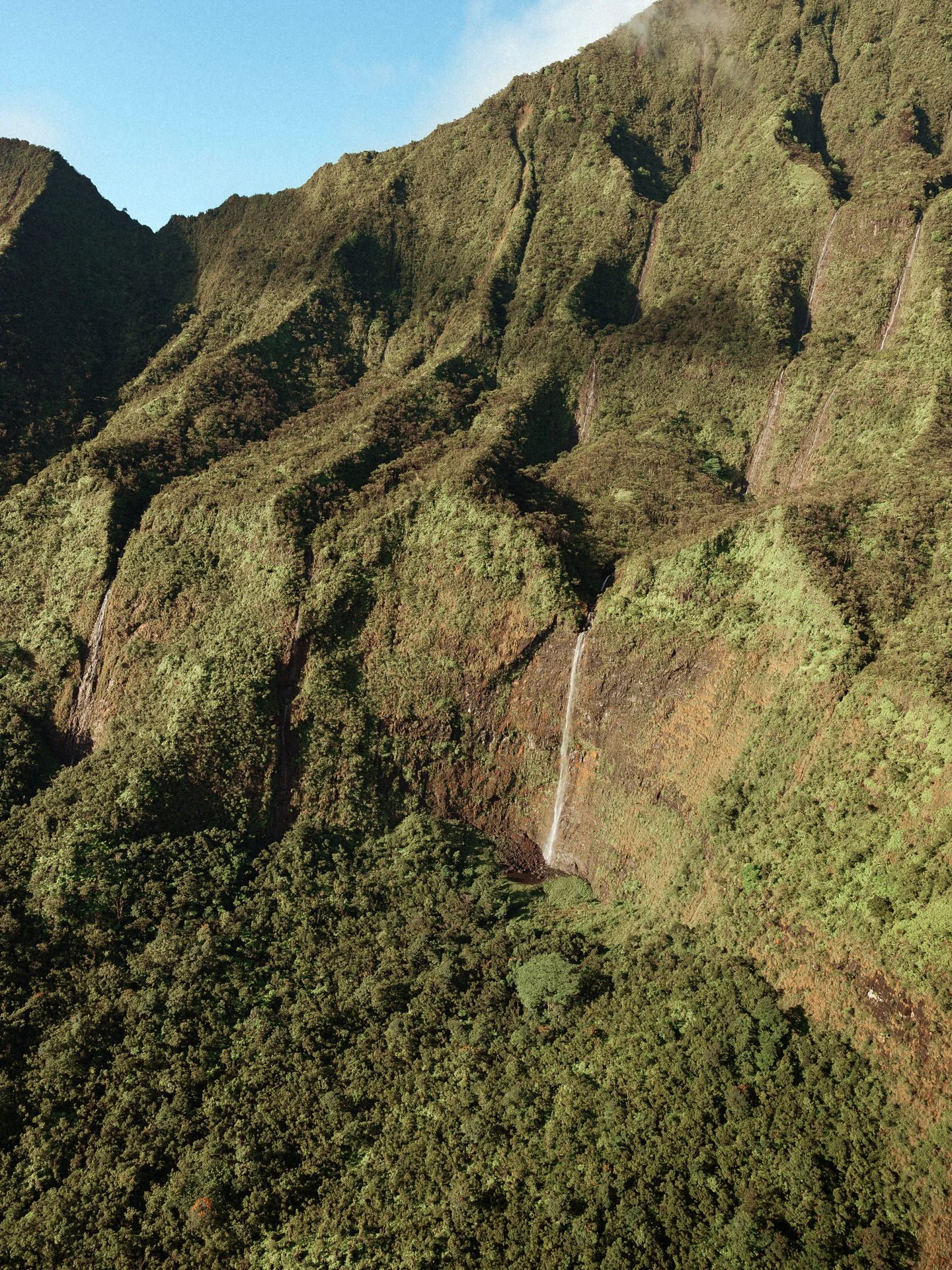 Aerial view of lush green mountainous landscape featuring multiple small waterfalls cascading down the slopes.
