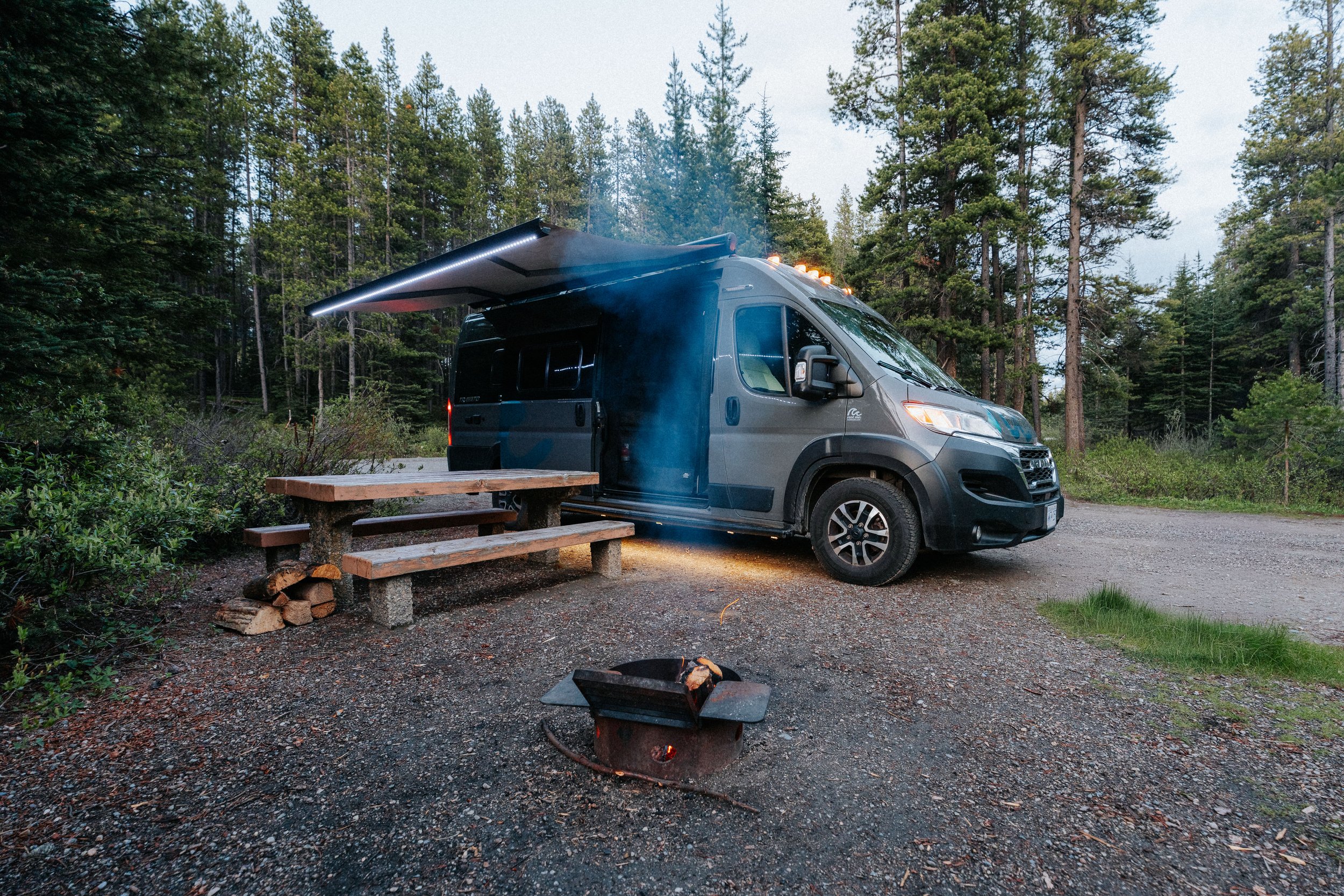 A gray camper van with a retractable awning parked in a forested area near a wooden picnic table, with a small campfire in the foreground under a cloudy sky.