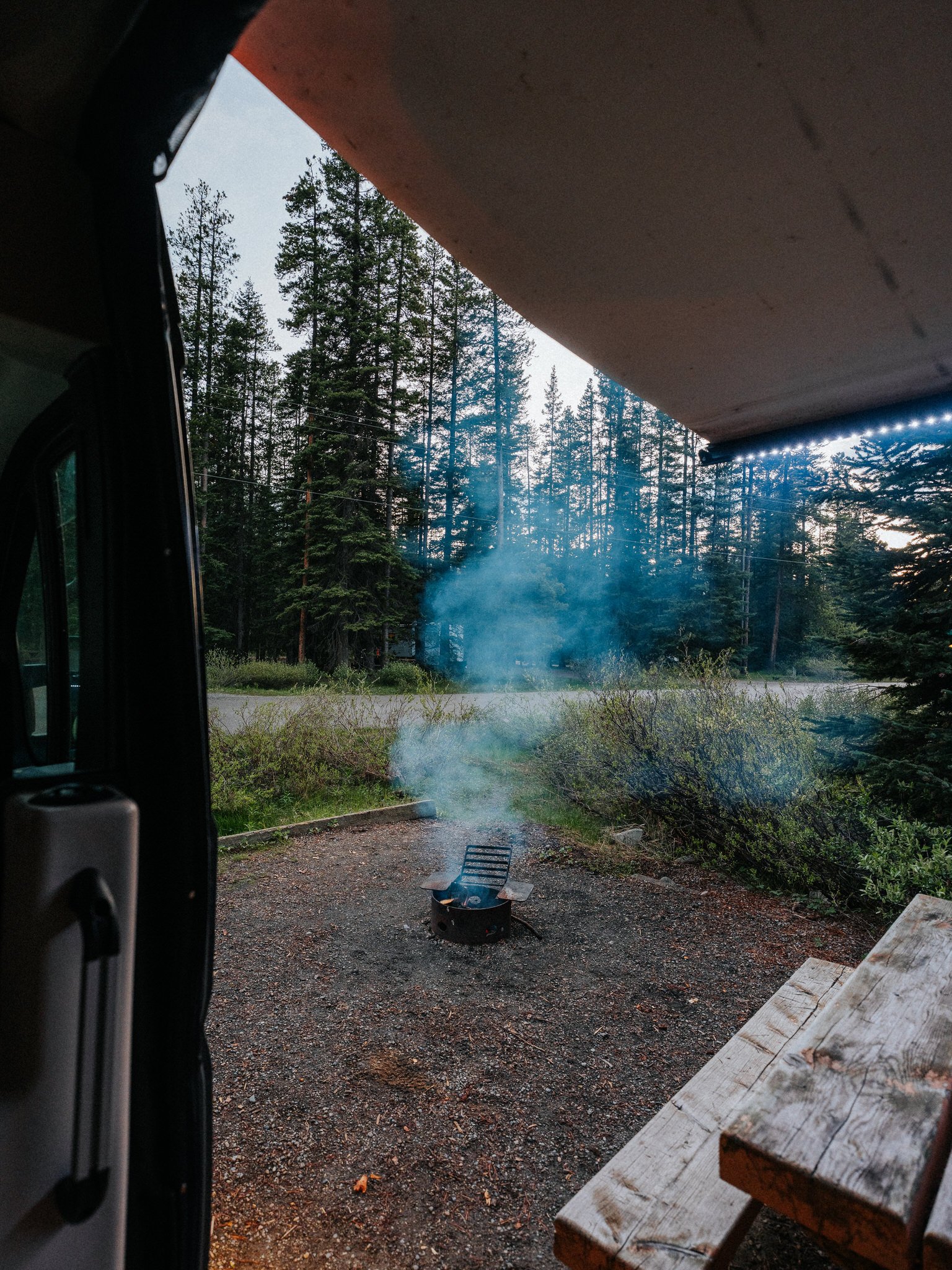 View from inside a vehicle showing an outdoor camping scene with a small campfire, smoke, a picnic table, and a forest of tall pine trees, with the sky above.