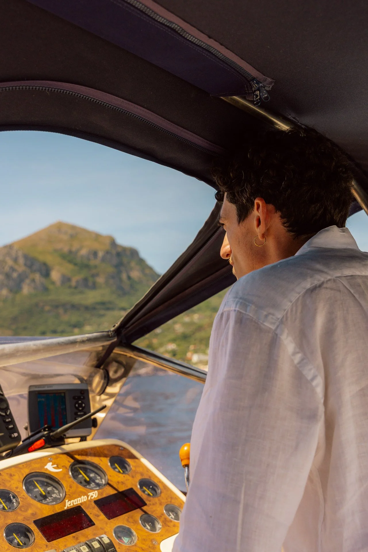 A person with dark curly hair and a hoop earring, dressed in a white shirt, is looking out from a boat’s cabin at a scenic view of a mountain and water with clear skies.