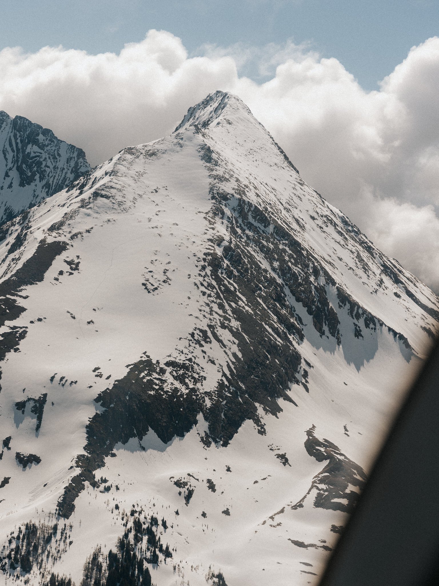 Snow-covered mountain peak with dark rocky patches and evergreen trees at base, under cloudy sky.
