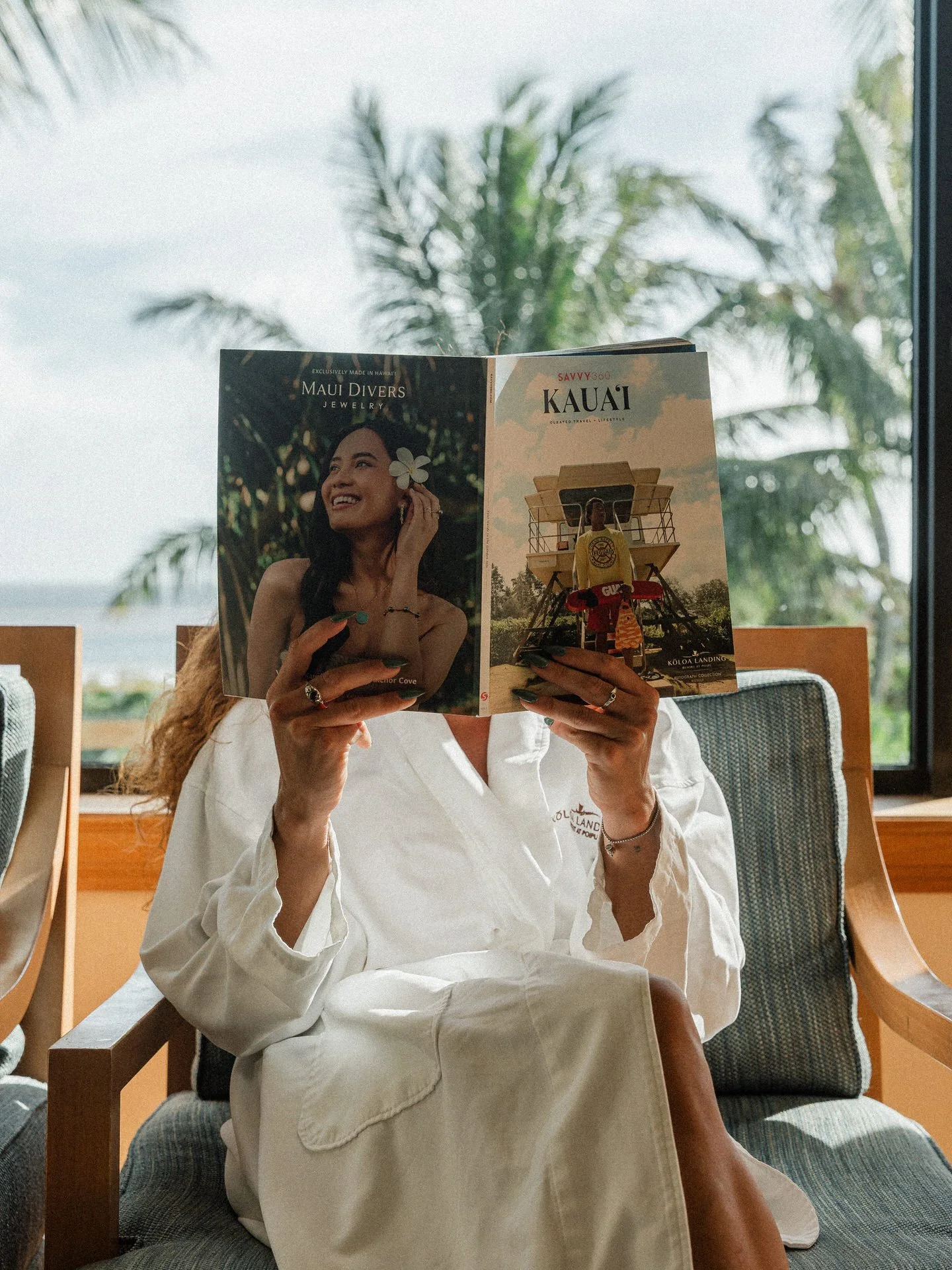 A woman sitting on a chair reading a travel magazine, with palm trees and a window view in the background.