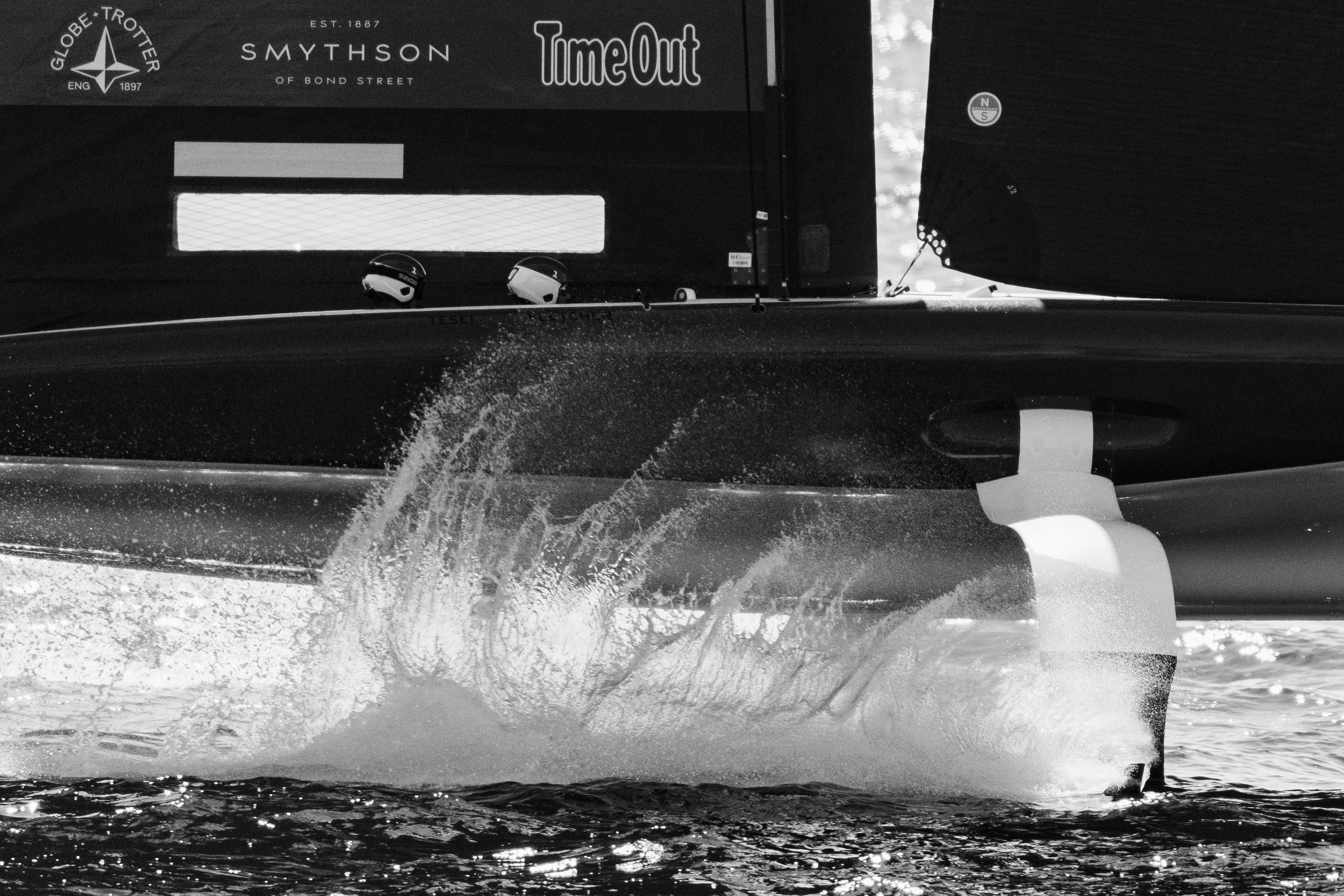 A black and white image of the back of a boat moving through water, creating a wake. visible on the boat are stickers and logos, including 'Globe Trottler,' 'Smythson,' and 'Time Out.' The boat's hull is partially visible at the bottom of the image.