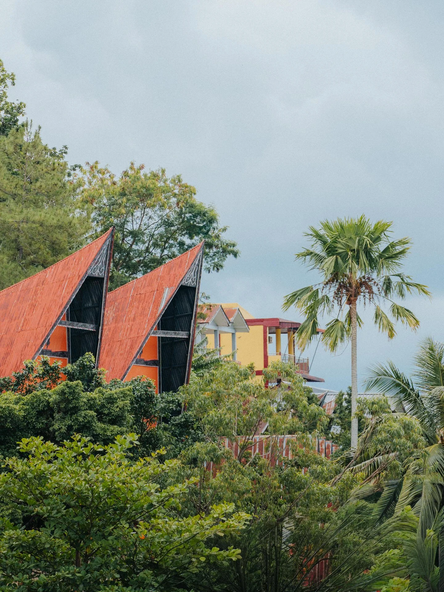 Tropical scene with modern buildings featuring red and yellow roofs, surrounded by dense green foliage and palm trees, under a cloudy sky.