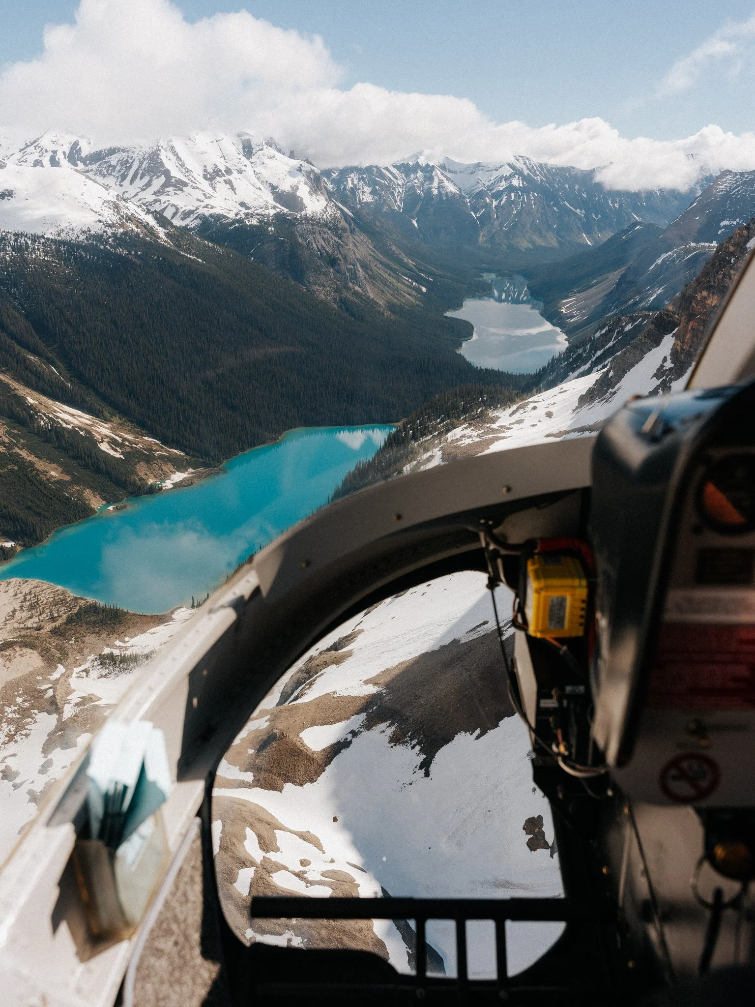 A view from inside a helicopter cockpit showing snow-covered mountains, a lake, and forests below, with part of the helicopter's control panel visible.