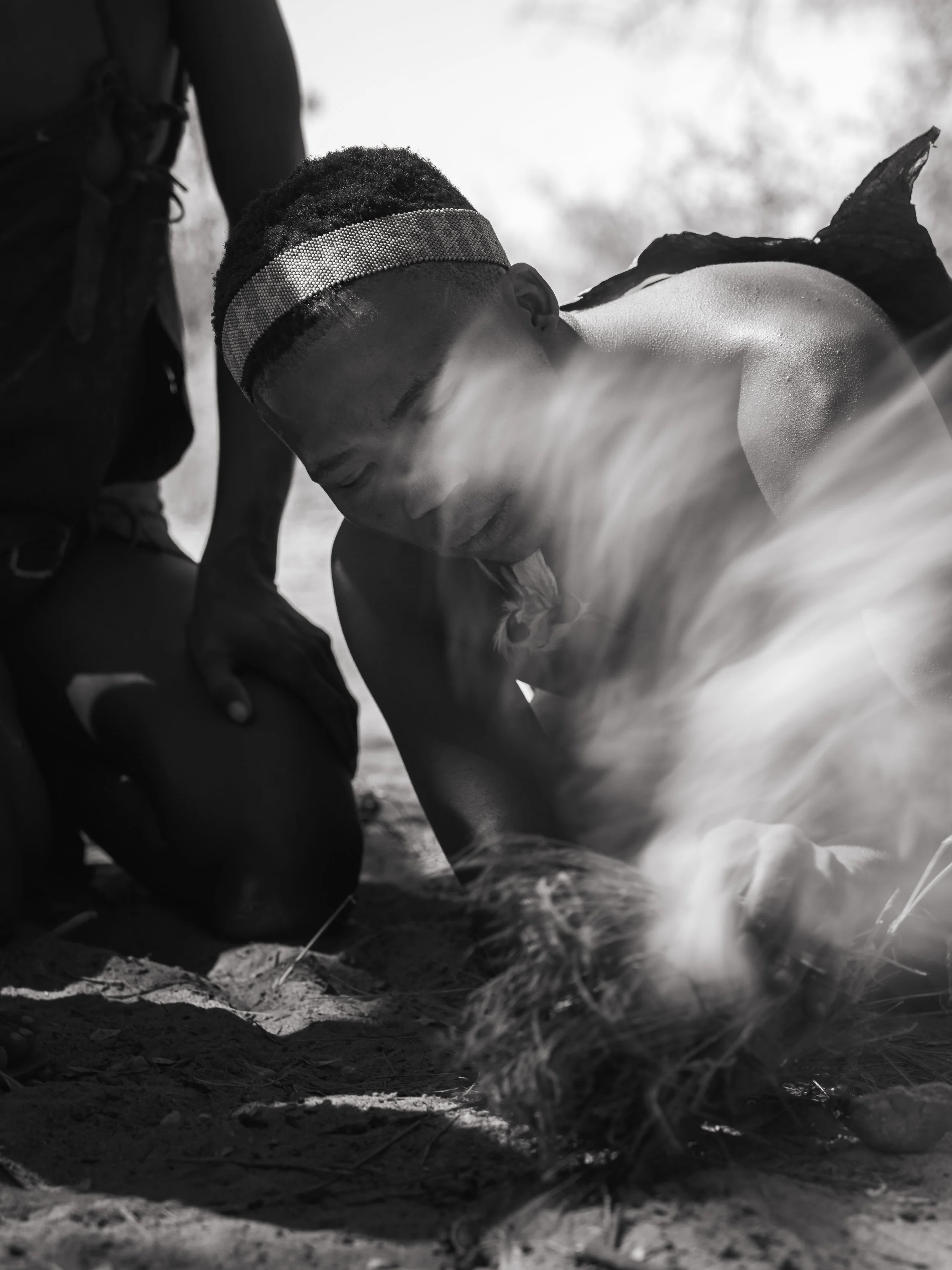 A black and white photograph of a person with a headband, lying on the ground in a natural outdoor setting. The person appears to be resting or unconscious, with their eyes closed and head tilted down.