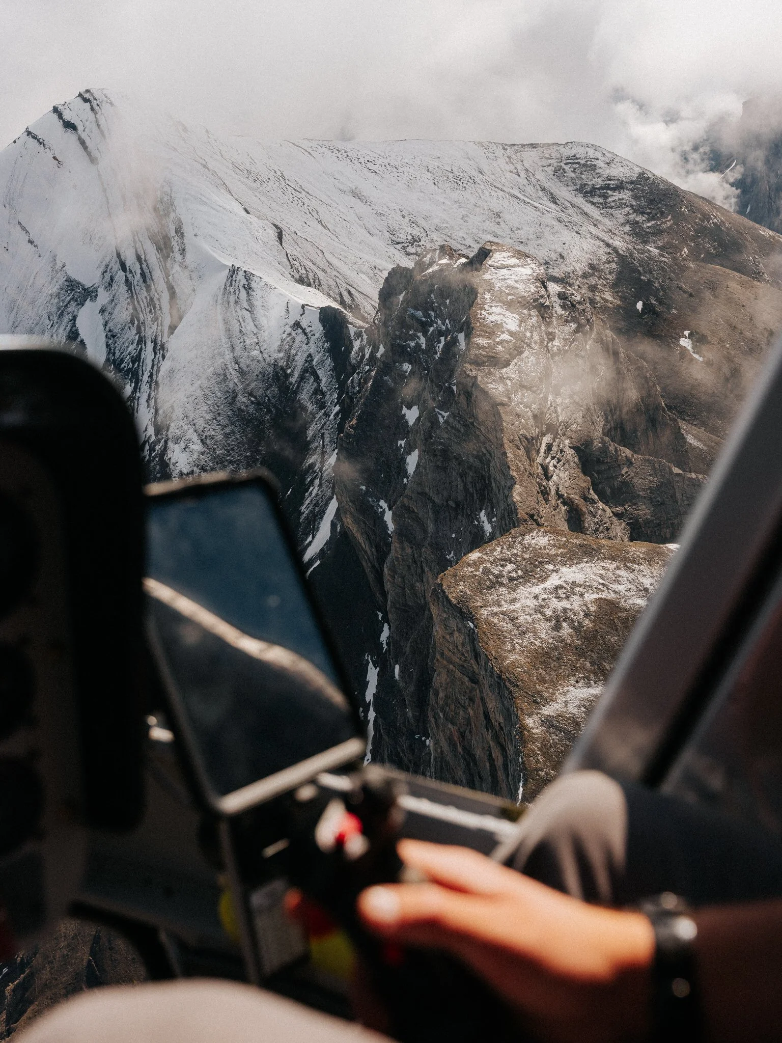Snow-covered mountain peaks seen from the window of an aircraft with a person's hand adjusting controls in the foreground.
