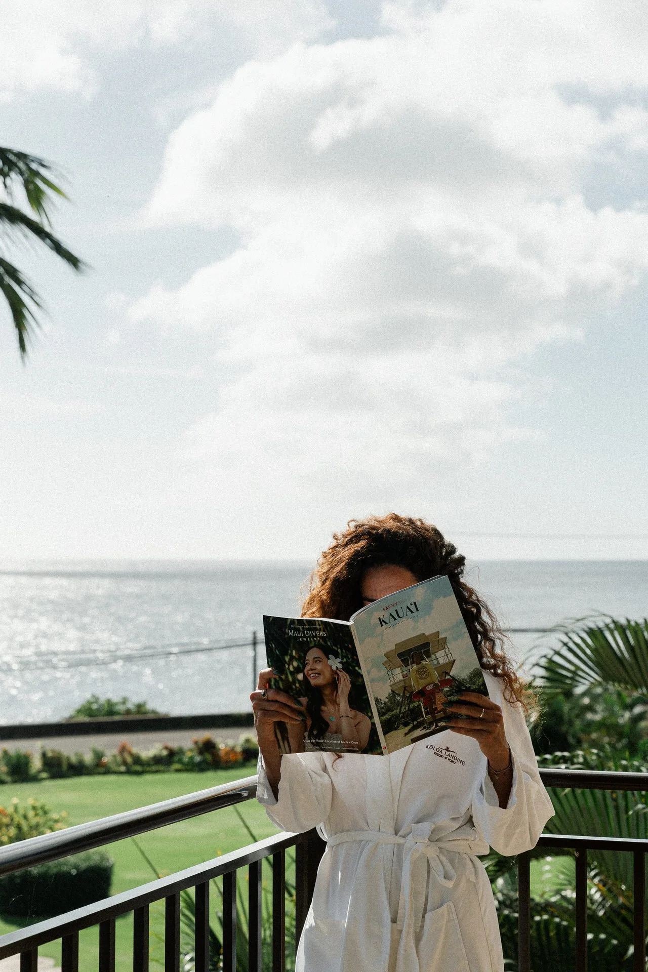 Woman in a white robe reading a magazine on a balcony overlooking the ocean, with palm trees and greenery around.