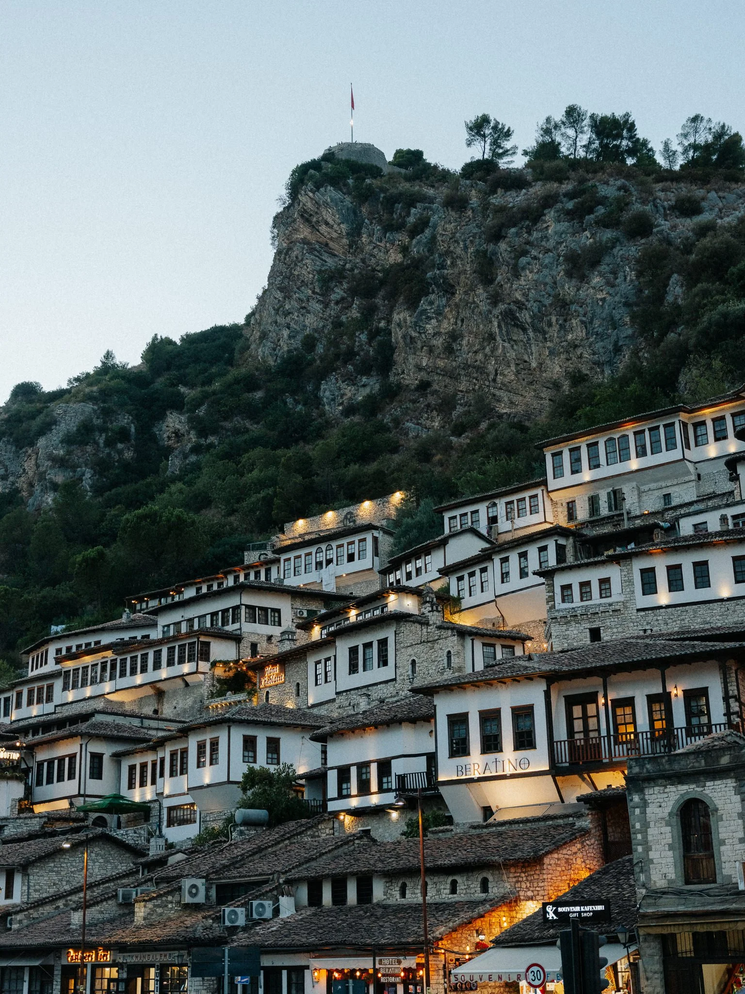 A hillside town with white buildings with black window frames and red-tiled roofs, set against a large rocky hill with sparse trees, during dusk, with signs of shops and hotels on the street level.