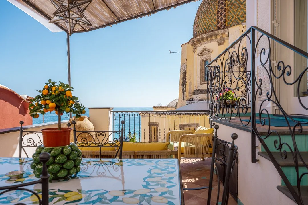 Balcony with outdoor furniture, a potted lemon tree, and a view of the sea and historic buildings under a sunny sky.